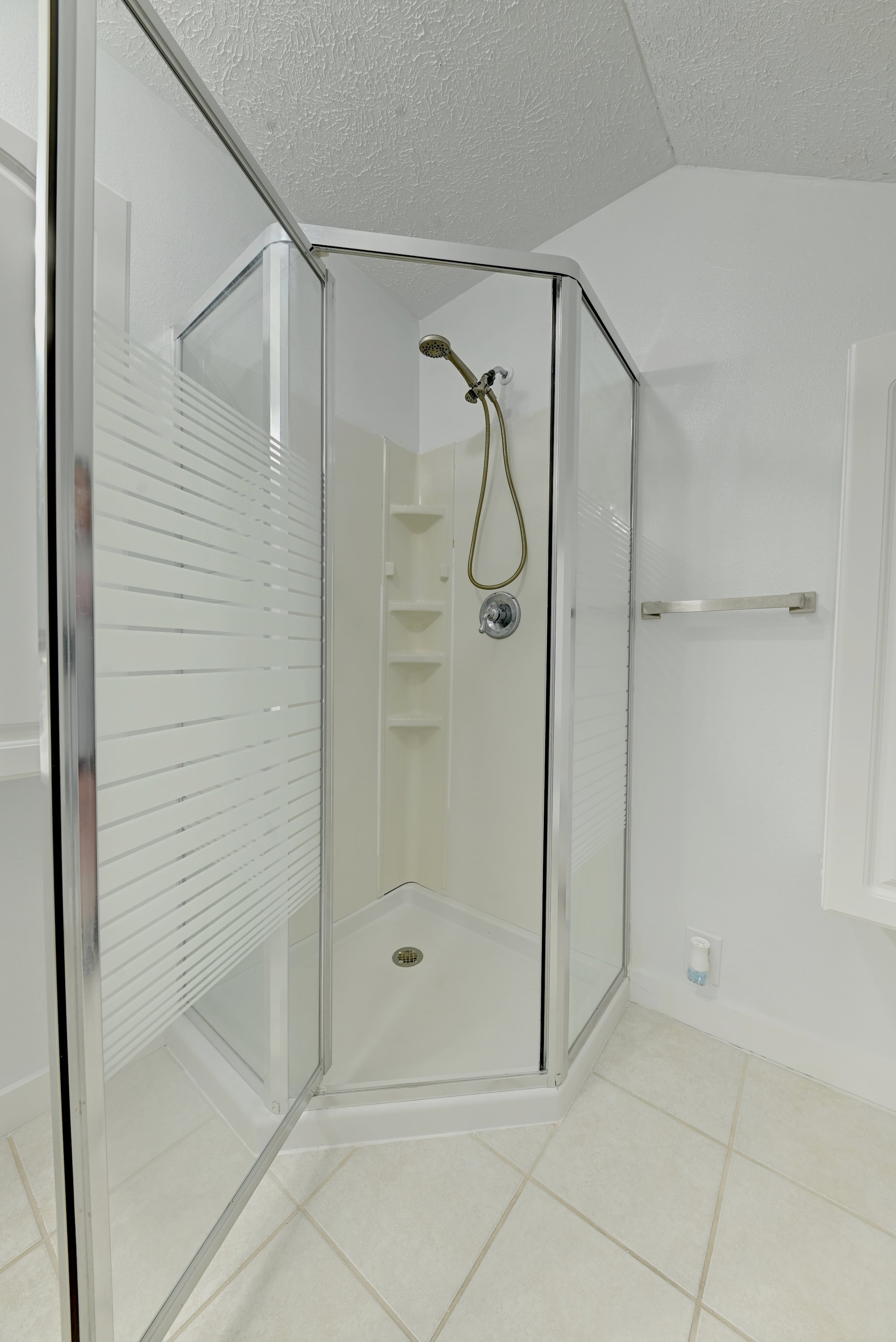A small enclosed shower with clear glass doors, a handheld showerhead, built-in shelves, and white tile walls in a bathroom.