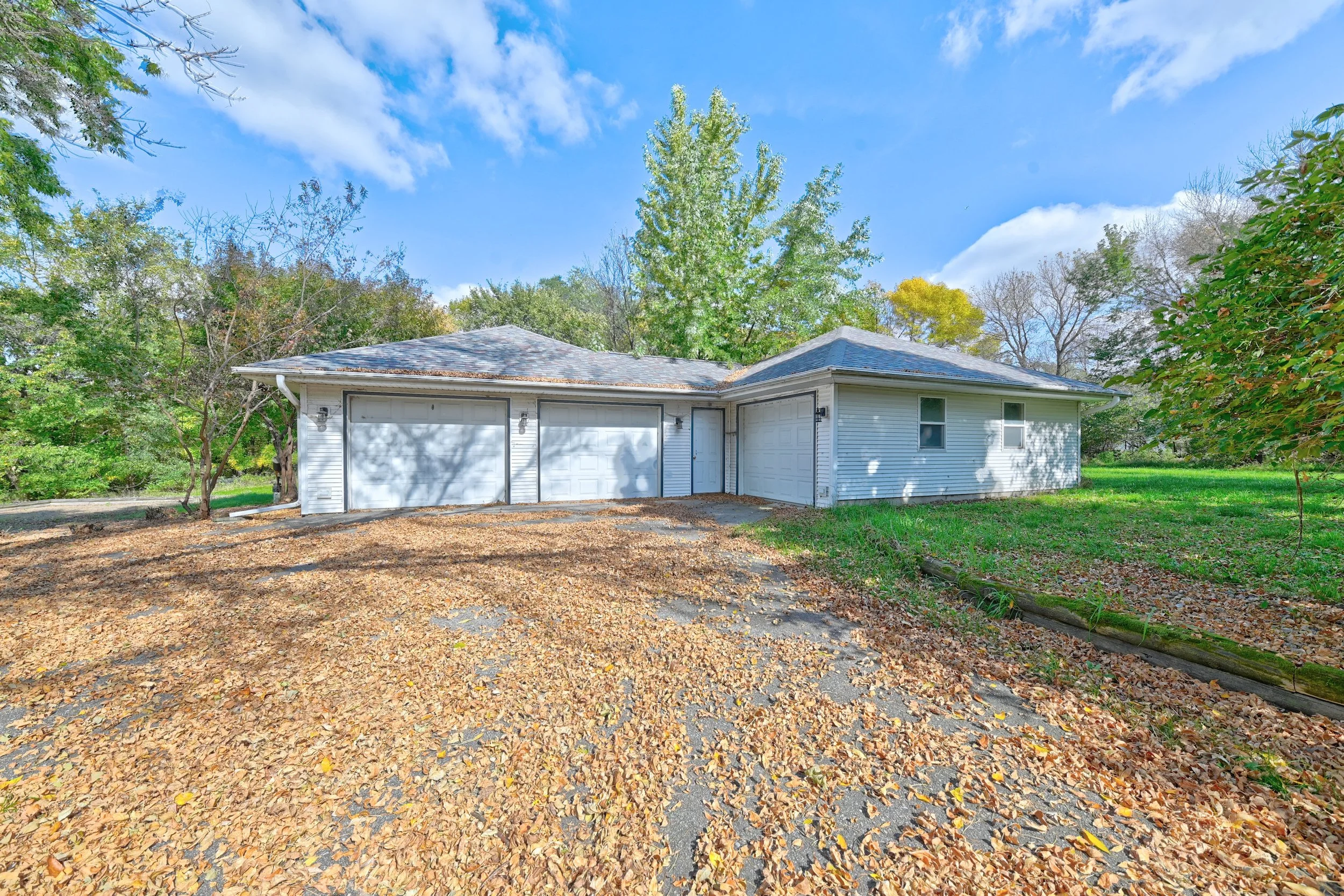 A white garage with two doors and a side door, surrounded by trees with green and some changing leaves, with fallen leaves on the ground and a blue sky with clouds.