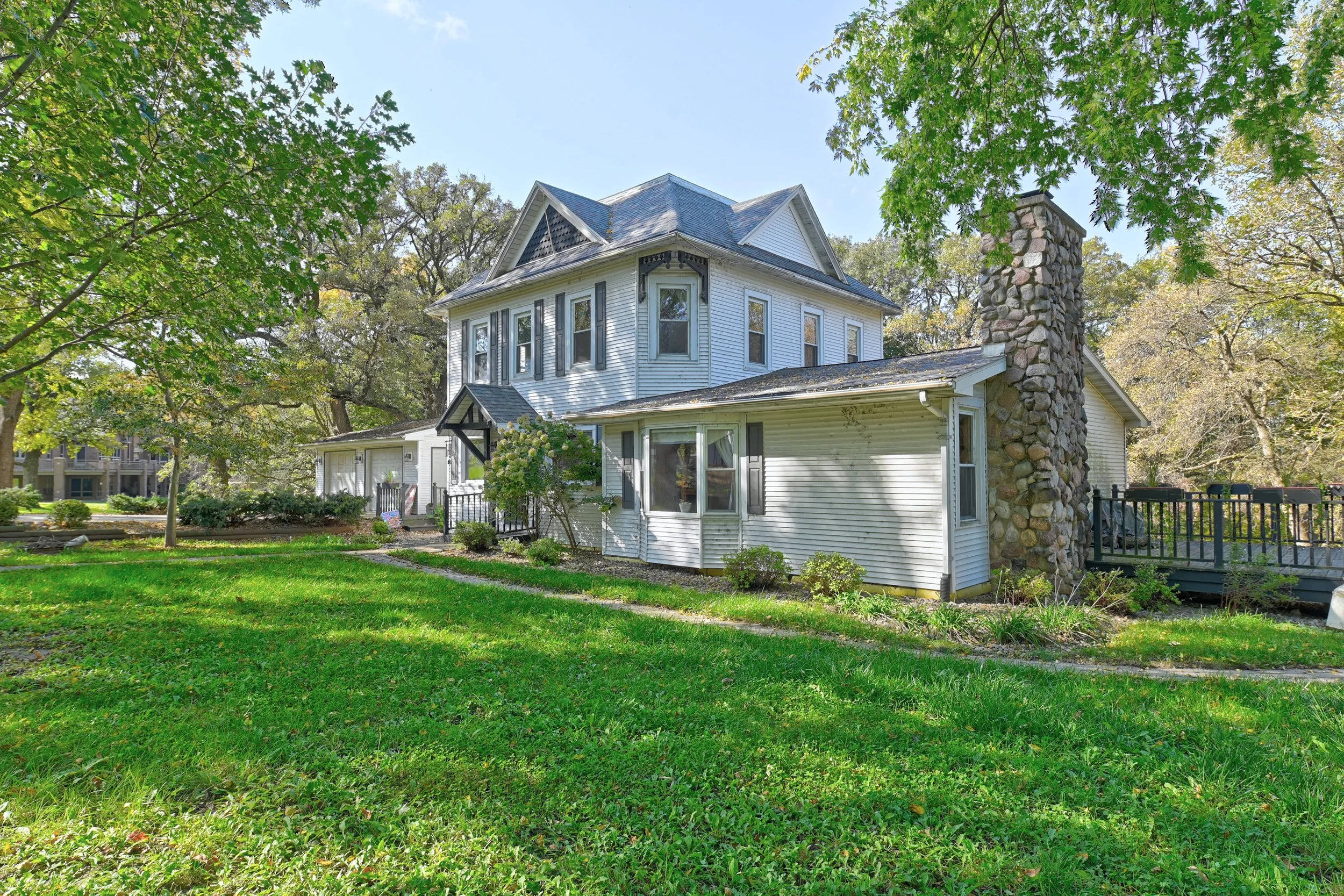 A white, two-story house with a stone chimney, surrounded by a green lawn and trees in the background on a sunny day.