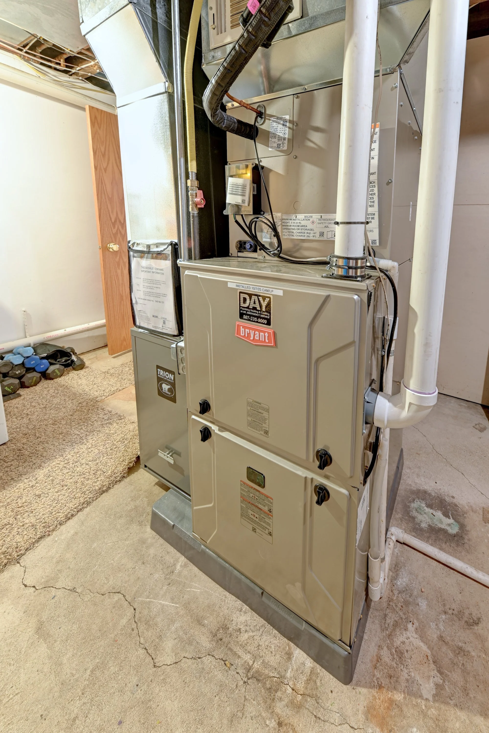 A commercial HVAC unit with a Bryant label, installed on a concrete floor, with a white PVC pipe connected to it, surrounded by a beige carpet and a small pile of dumbbells in the background.