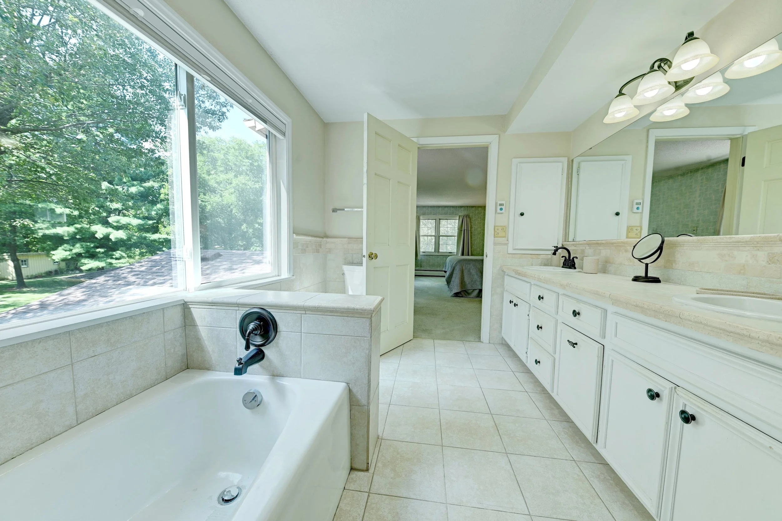 Bathroom with large window showing green trees outside, white vanity with multiple drawers, large mirror, bathroom sink with black faucet, toilet, and bedroom visible through open door.