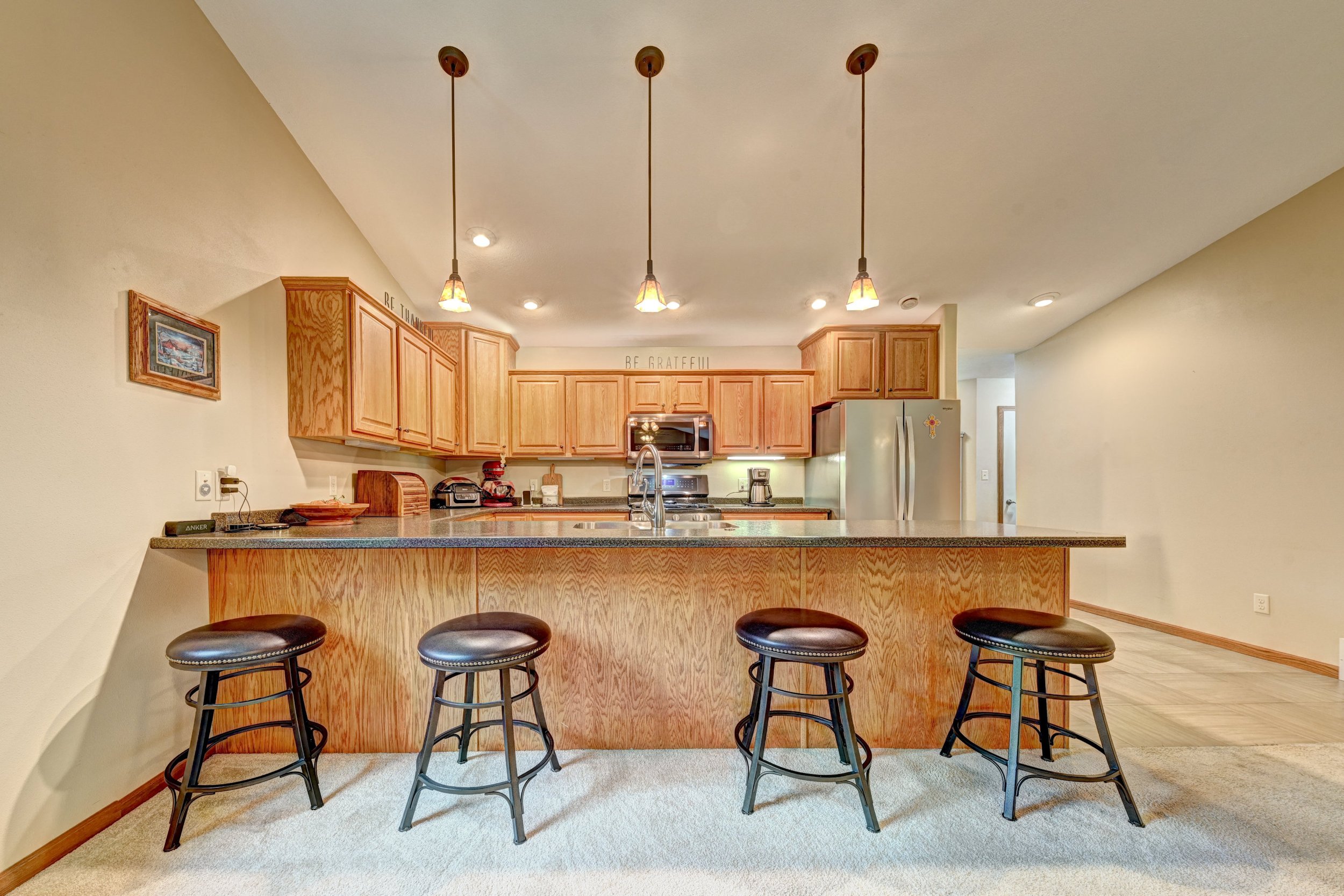 A kitchen with wooden cabinets, a granite countertop, and three pendant lights hanging above a breakfast bar with four black leather and metal bar stools. There are appliances and decor on the countertop, including a microwave, toaster, and a clock.