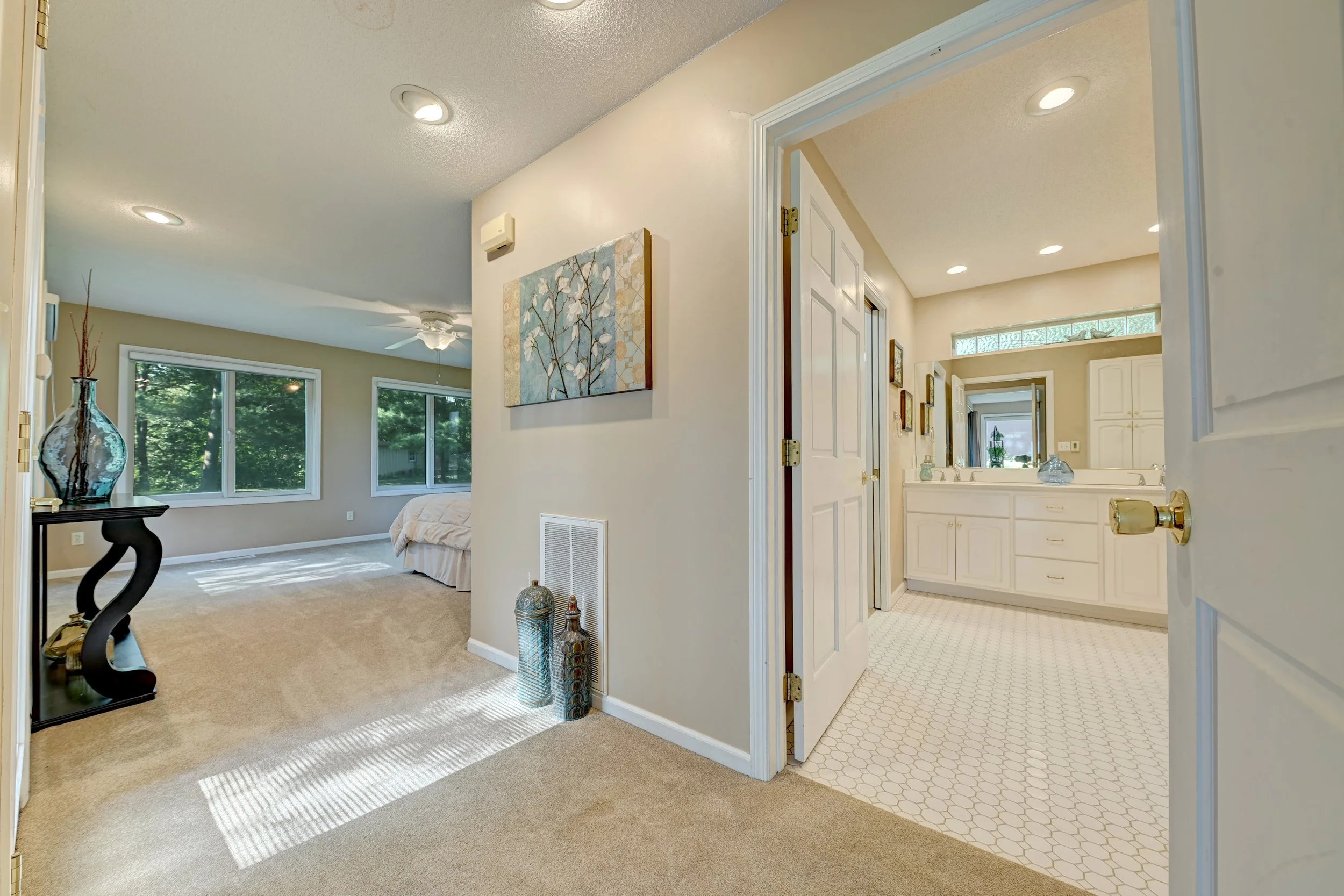 View of a cozy living room with beige carpet, large windows with trees outside, artwork on the wall, and a small decorative table with vases. The bathroom with white cabinets and a vanity mirror is visible through an open door.