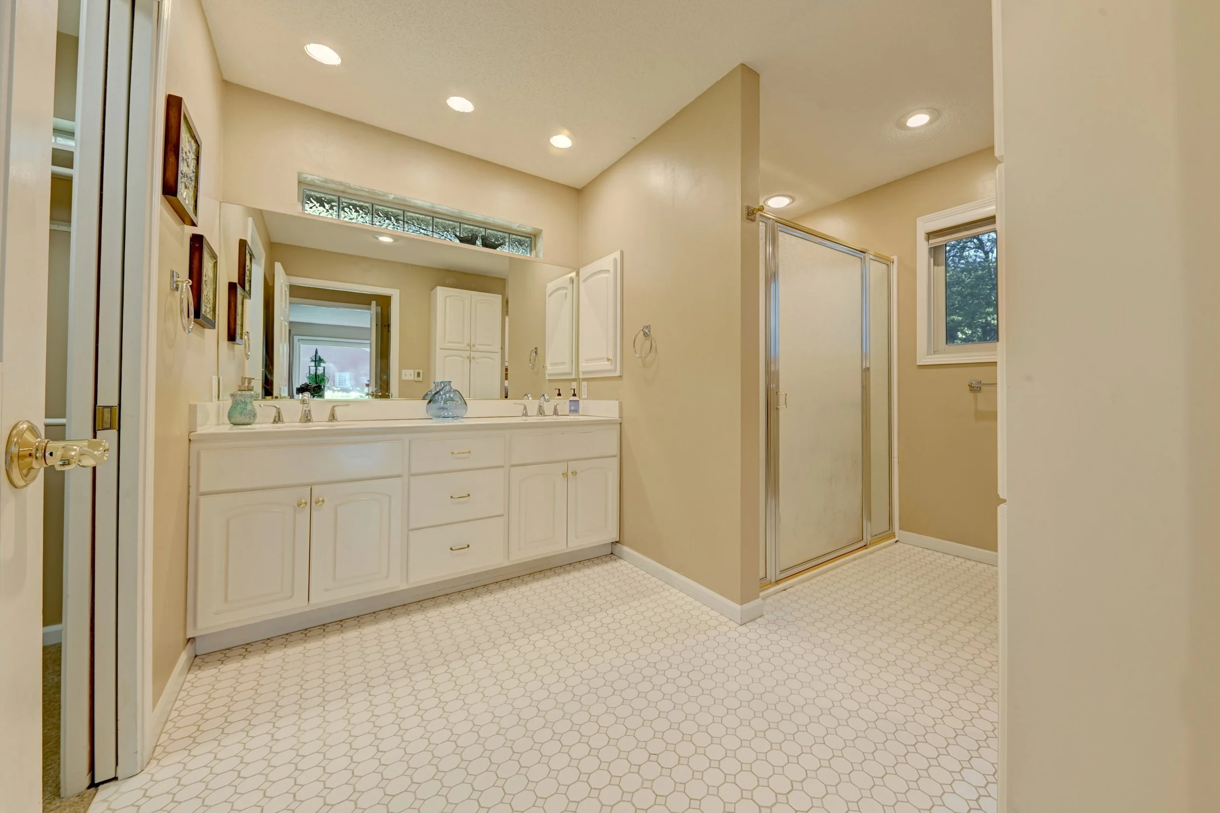 Bathroom with white vanity, large mirror, glass vase, beige walls, small window, shower with sliding door, and hexagonal pattern tile flooring.