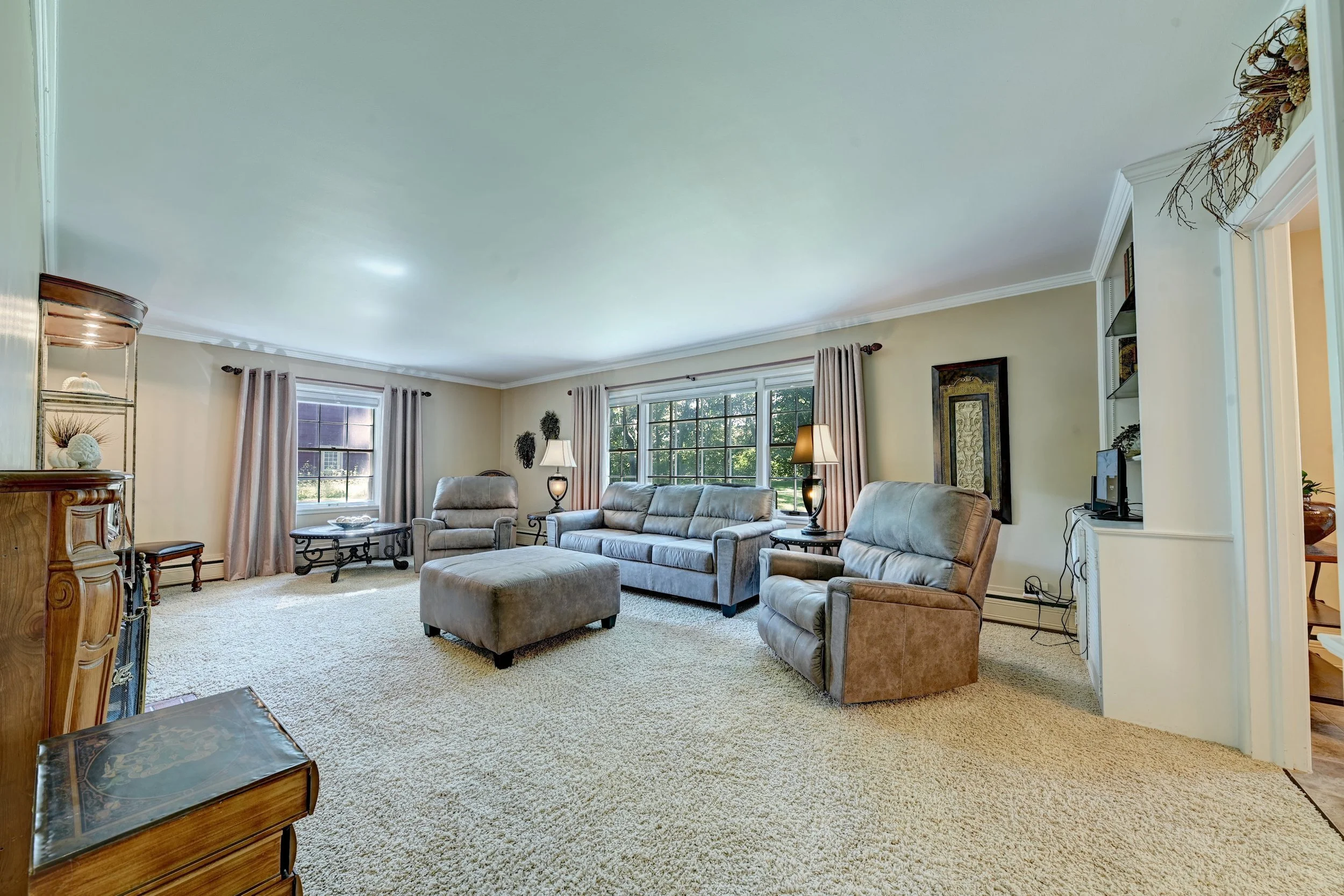 Living room with beige walls, large windows with curtains, a beige carpet, beige couches, end tables with lamps, a decorative wall art, and a corner bookshelf with decorative items.