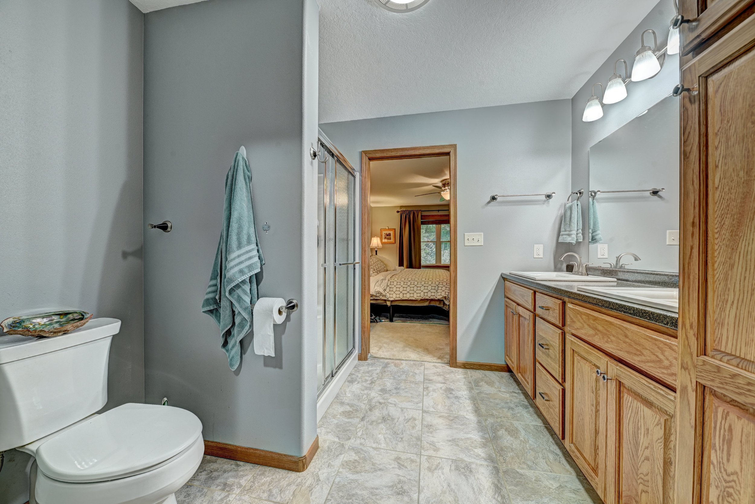 A bathroom with light gray walls, a toilet in the corner, a towel hanging on a hook, a shower stall, a double sink vanity with wooden cabinets, wall-mounted mirrors, and bright lighting above the mirrors. A doorway leads to a bedroom visible in the b