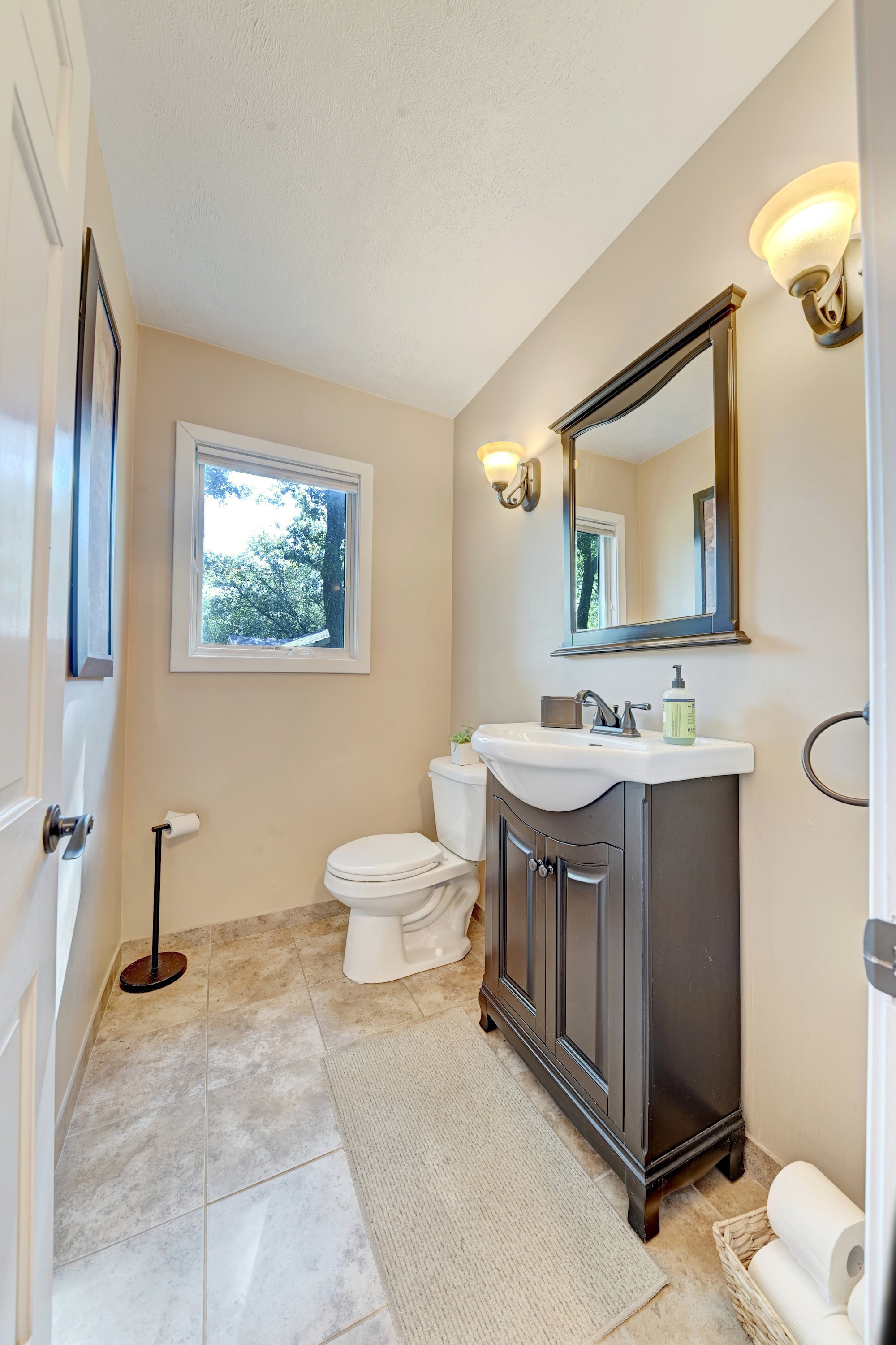 A bathroom with beige walls, a window showing trees outside, a white toilet, a brown-finished sink vanity with mirror, wall-mounted light fixtures, and a floor mat with toilet paper in a basket.