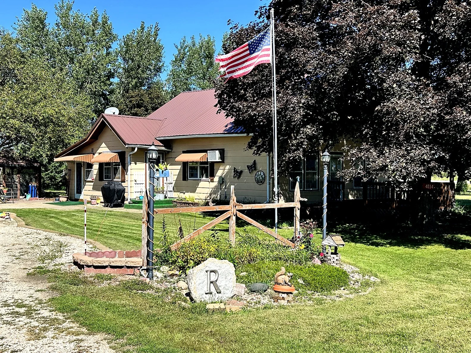 A house with a red metal roof, beige siding, and small front porch flanked by American flag on a tall pole. There are trees with green and dark purple leaves and well-maintained lawn and garden with a white stone marked 'R', flower bed, and garden or