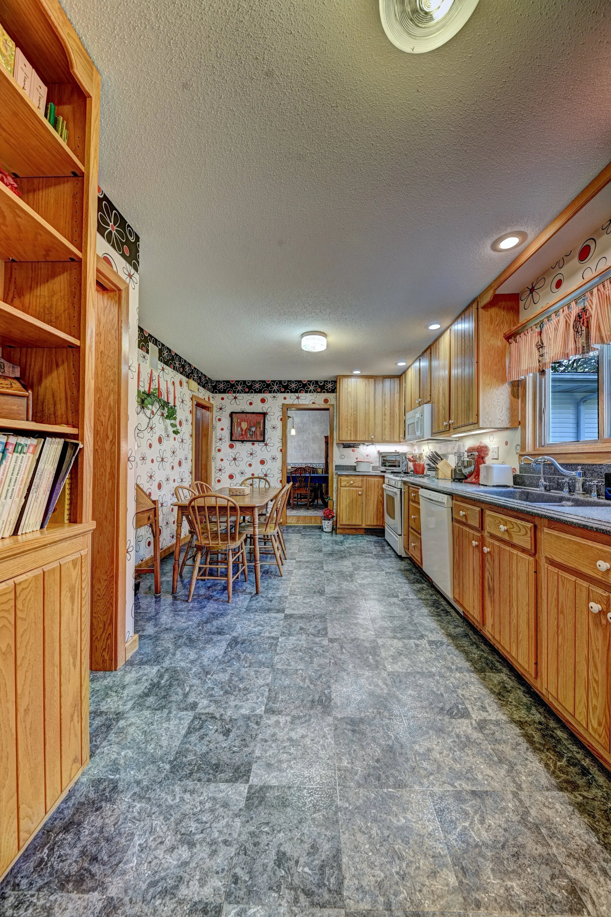 A kitchen with wooden cabinets, a dining table with chairs, and a wallpapered wall with floral patterns. The floor is tiled with stone-like vinyl, and the ceiling has recessed lighting and a ceiling fixture.