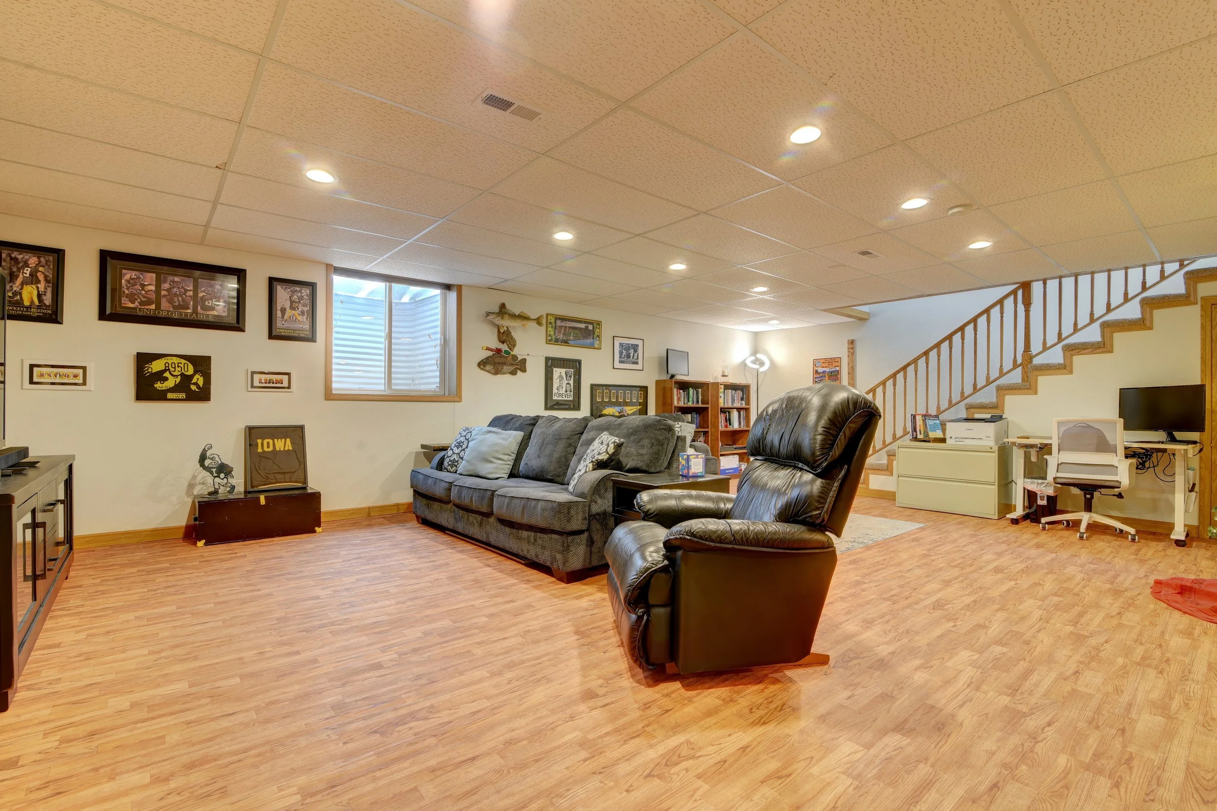 Living room with wood flooring, beige ceiling with recessed lighting, gray sofa with pillows, black leather recliner, framed pictures and sports memorabilia on the wall, small window, staircase with wooden railing, desk with a computer and printer, b
