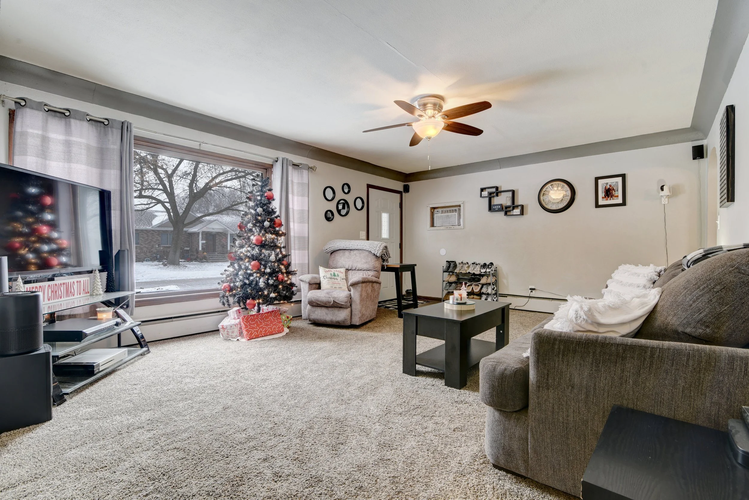 Living room decorated for Christmas with a Christmas tree, wrapped presents, and holiday decorations on the TV stand. There is a recliner chair, a sofa with white pillows, a black coffee table, and a shoe rack near a small window. The room has a larg