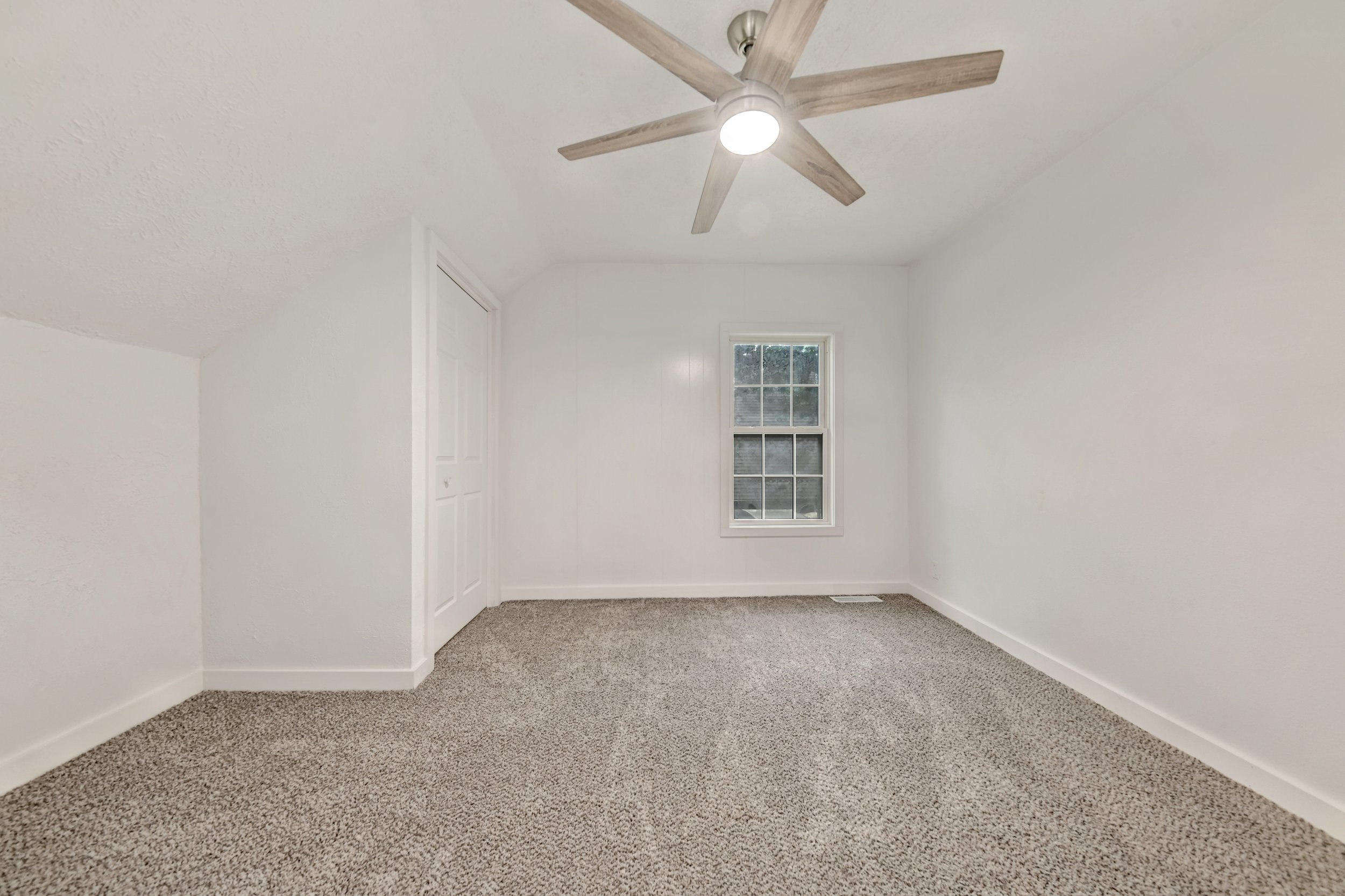 Empty room with beige carpet, white walls, a ceiling fan with light, a window, and a door.