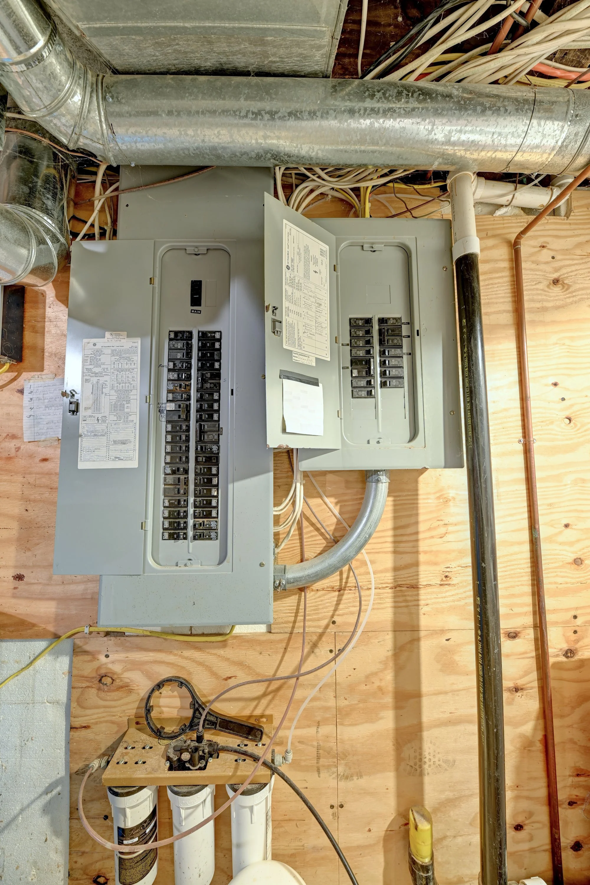 An electrical panel with open circuit breakers and wiring, mounted on a wooden wall in a basement or utility room, with plumbing pipes and tools nearby.