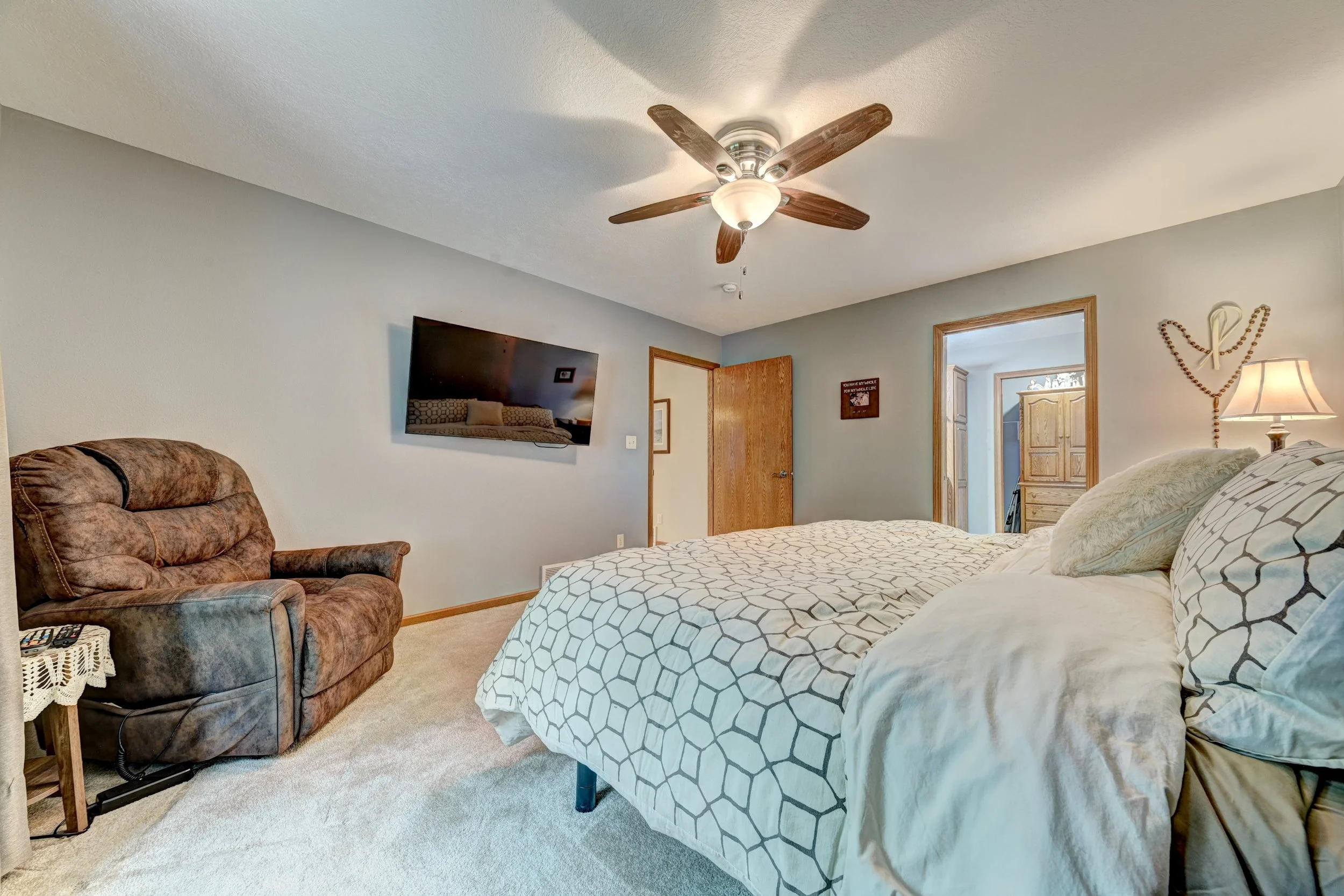 Bedroom with a bed, patterned bedding, bedside lamp, wall decor, a TV, a brown armchair, and an open door leading to an adjacent room.