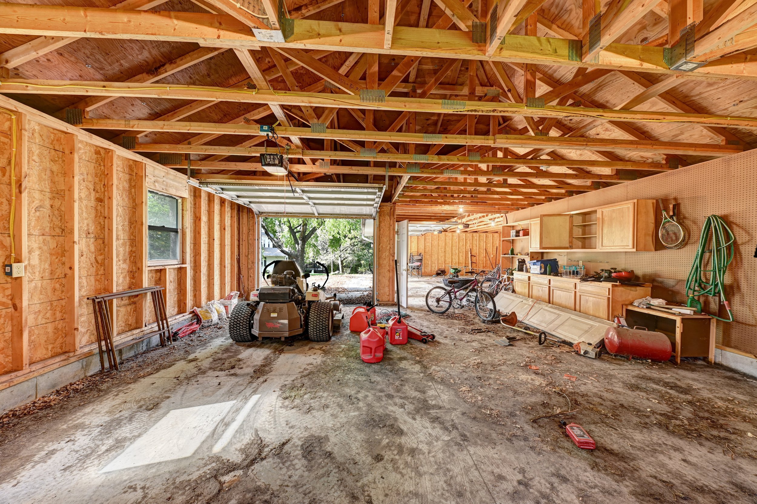 The interior of a garage under construction with exposed wooden framing and unfinished walls. There are bicycles, a lawnmower, and various tools and equipment scattered around. The open garage door shows trees and a yard outside.