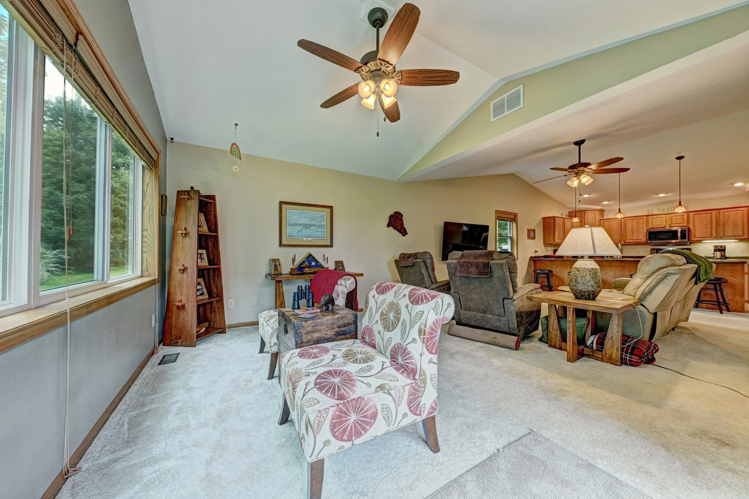Living room with large window, ceiling fans, and furniture including armchairs, sofa, and a coffee table, with an open kitchen area in the background.