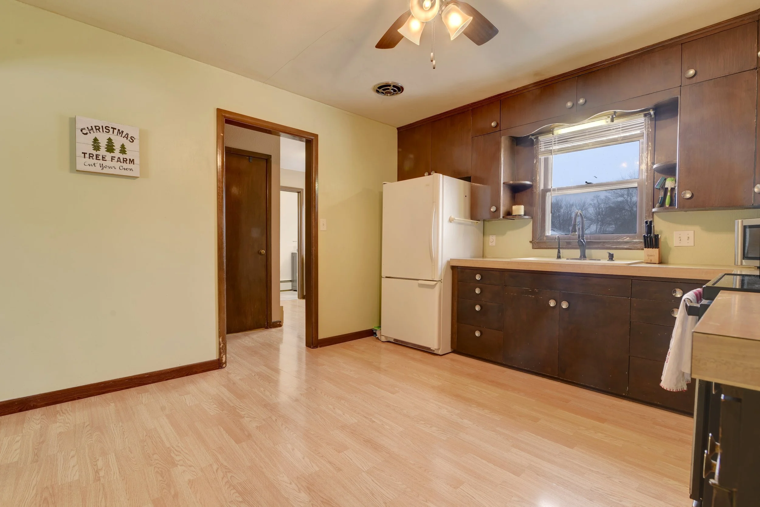 A kitchen with light wood flooring, dark wood cabinets, a white refrigerator, and a window above the sink showing trees outside.