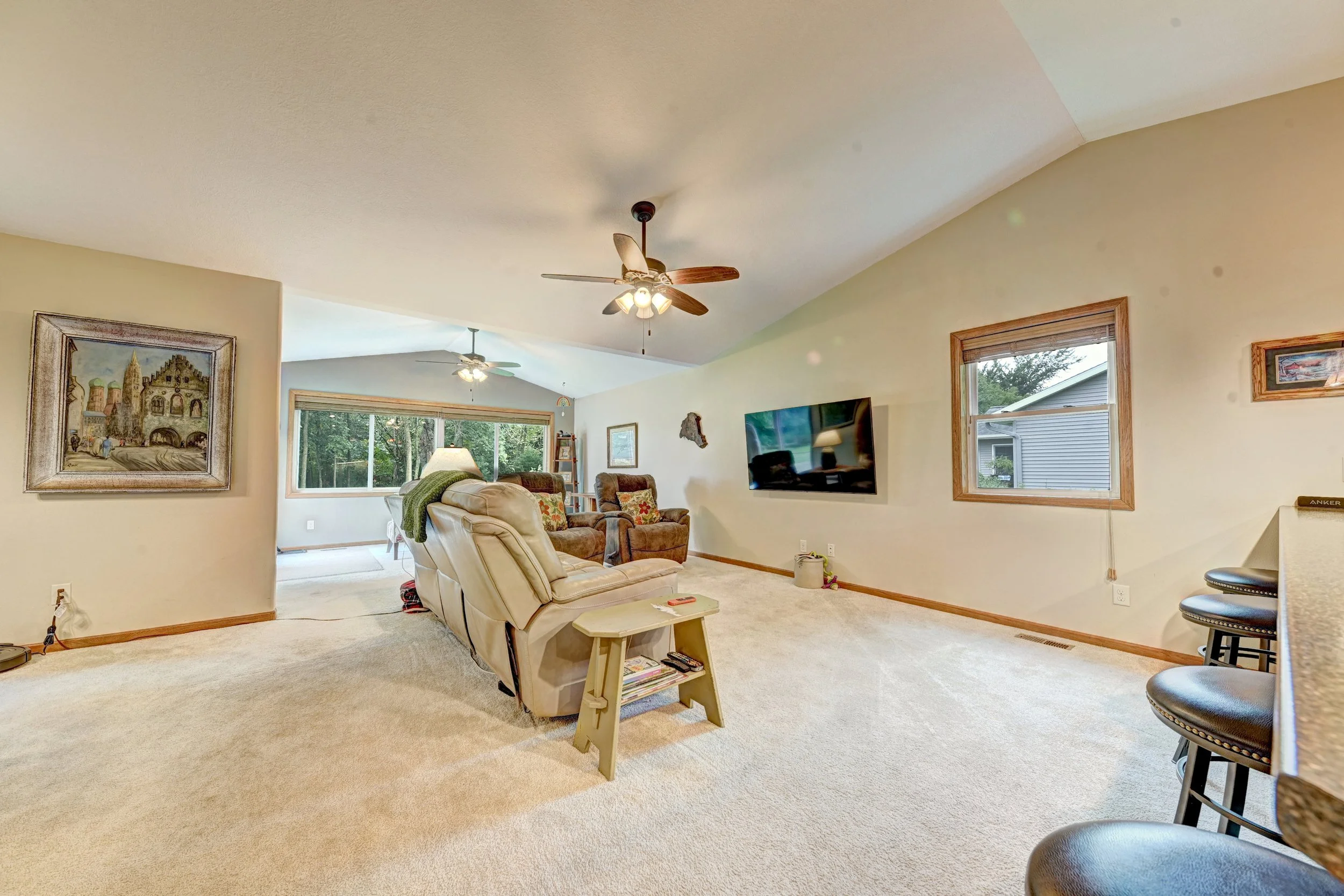 Interior view of a living room with beige carpet, reclining chairs, wall-mounted TV, windows showing outdoor trees, ceiling fans, and artwork on the walls.