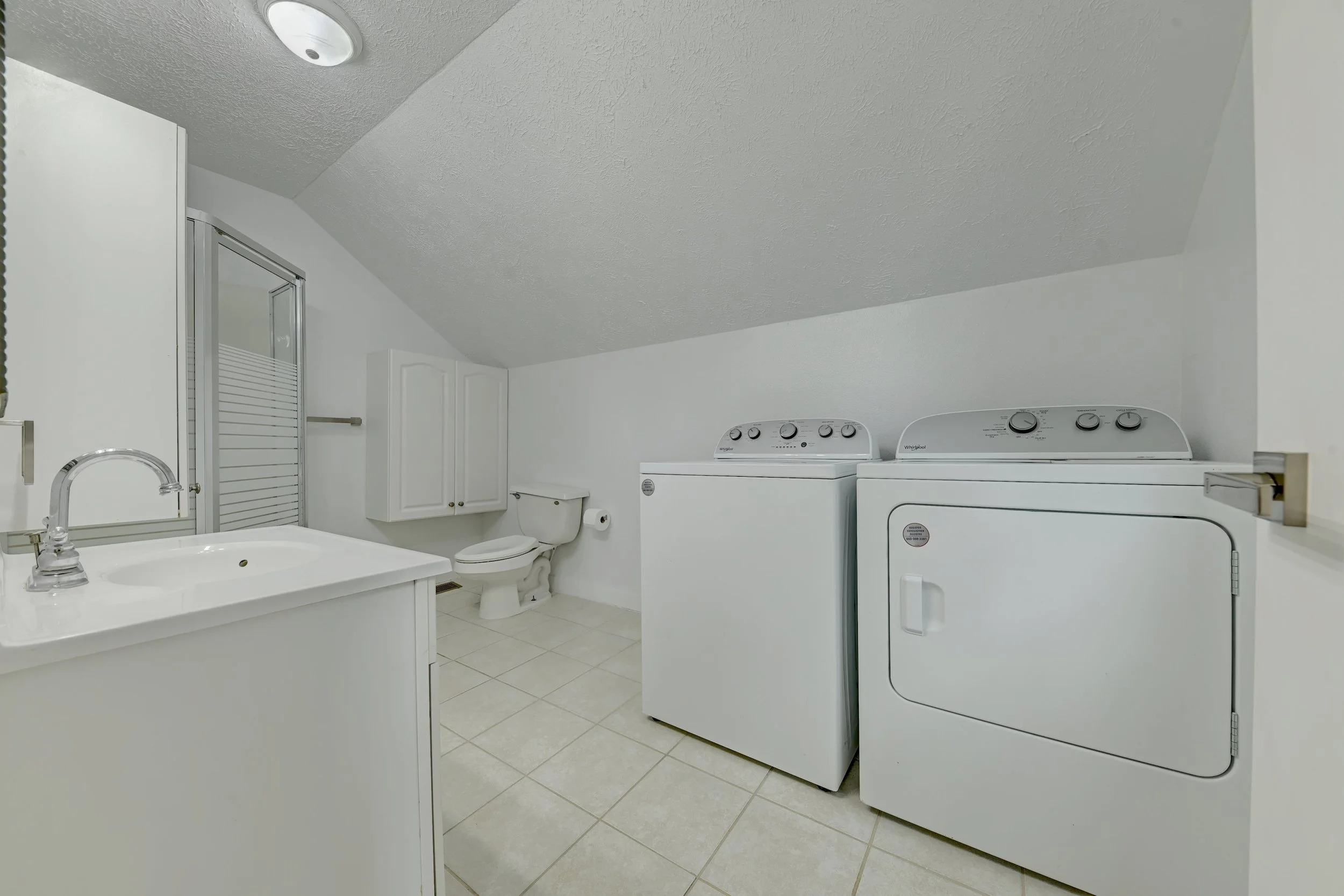 A laundry room with white appliances, including a washing machine and dryer, white cabinets, a toilet, a small sink, and a ceiling light.