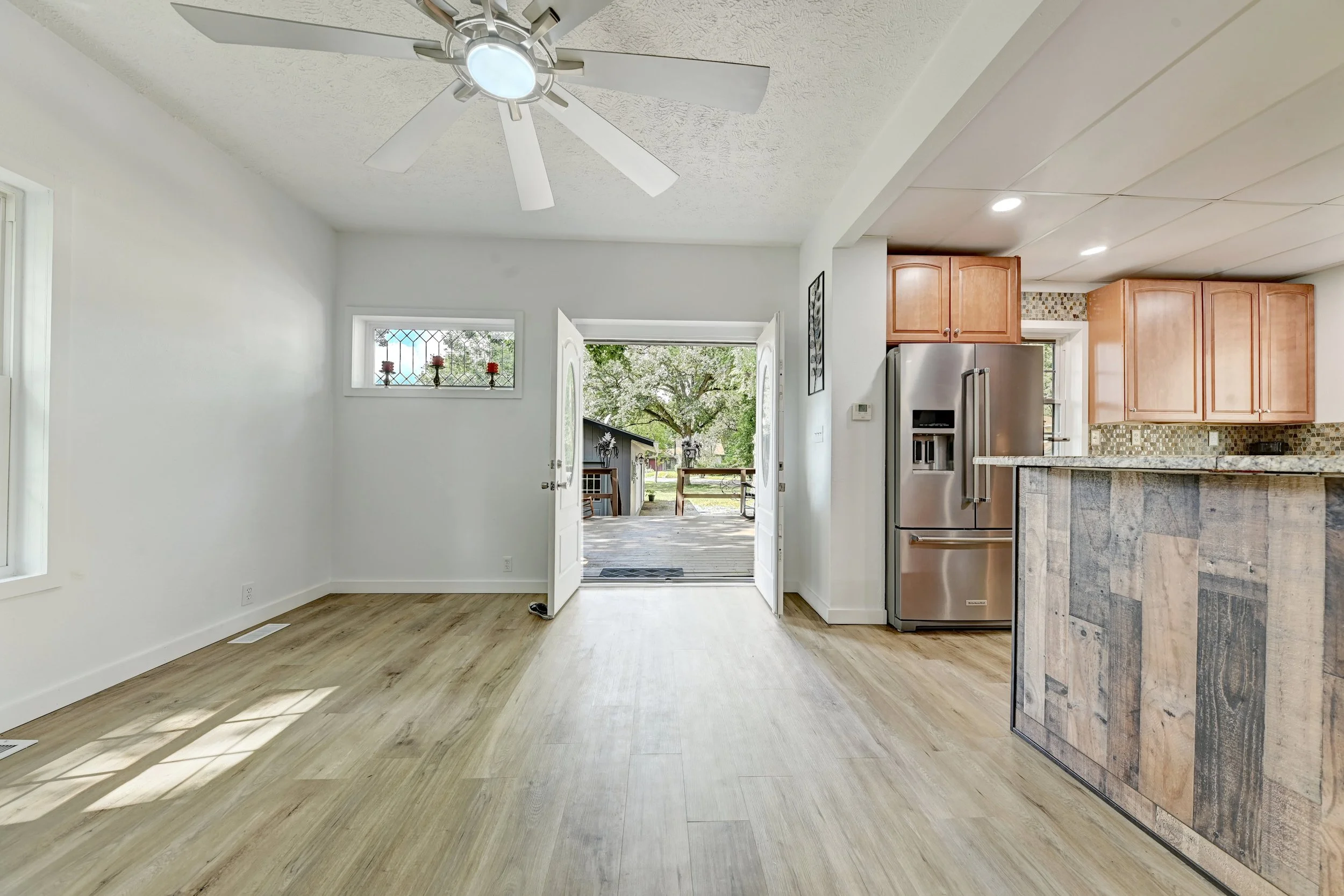 Interior view of a kitchen and dining area with white walls, wood flooring, a ceiling fan, and an open door leading outside to a porch with trees.