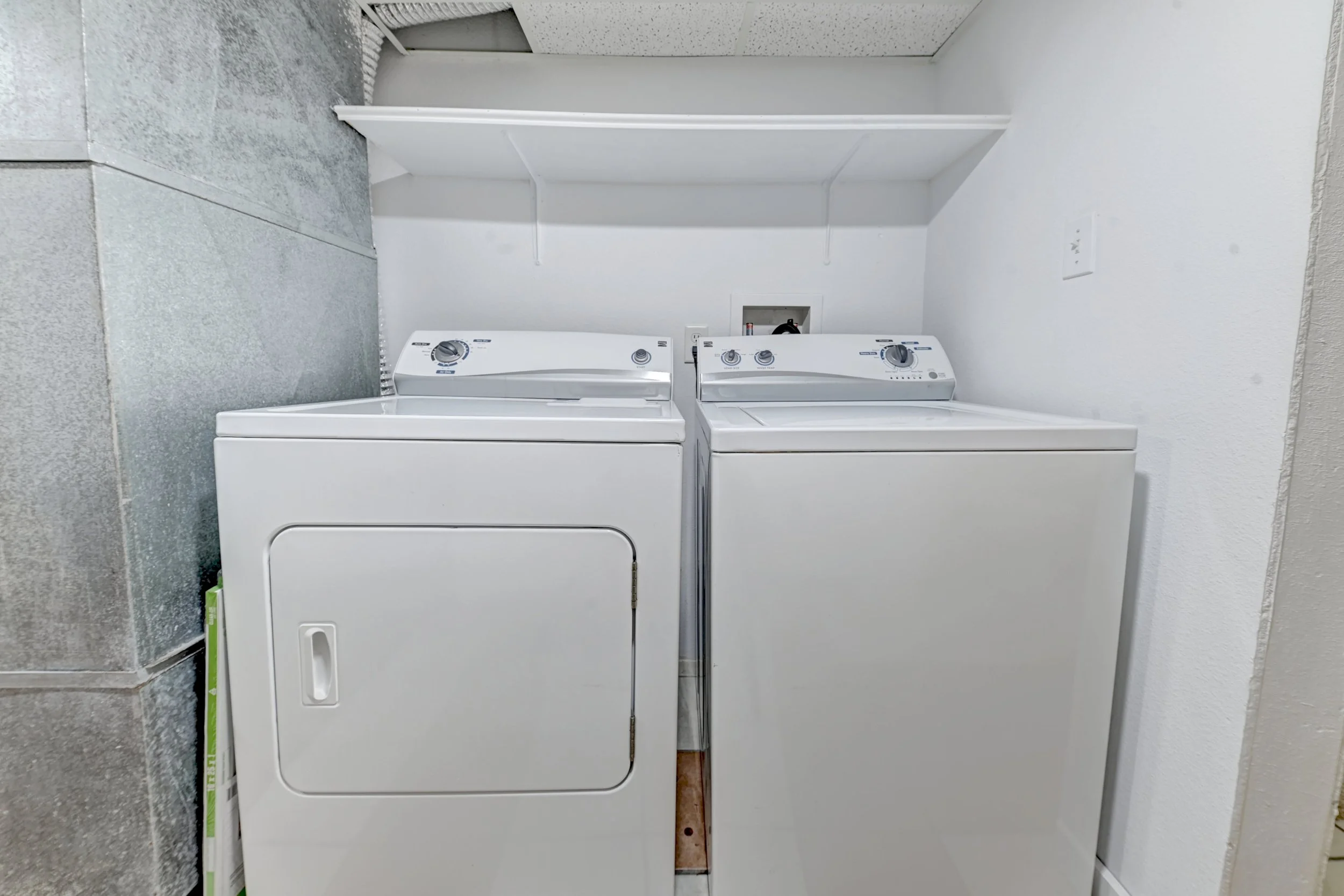 A laundry room with a white washing machine and dryer side by side against a white wall, with a white shelf above them and a small electrical panel on the wall.