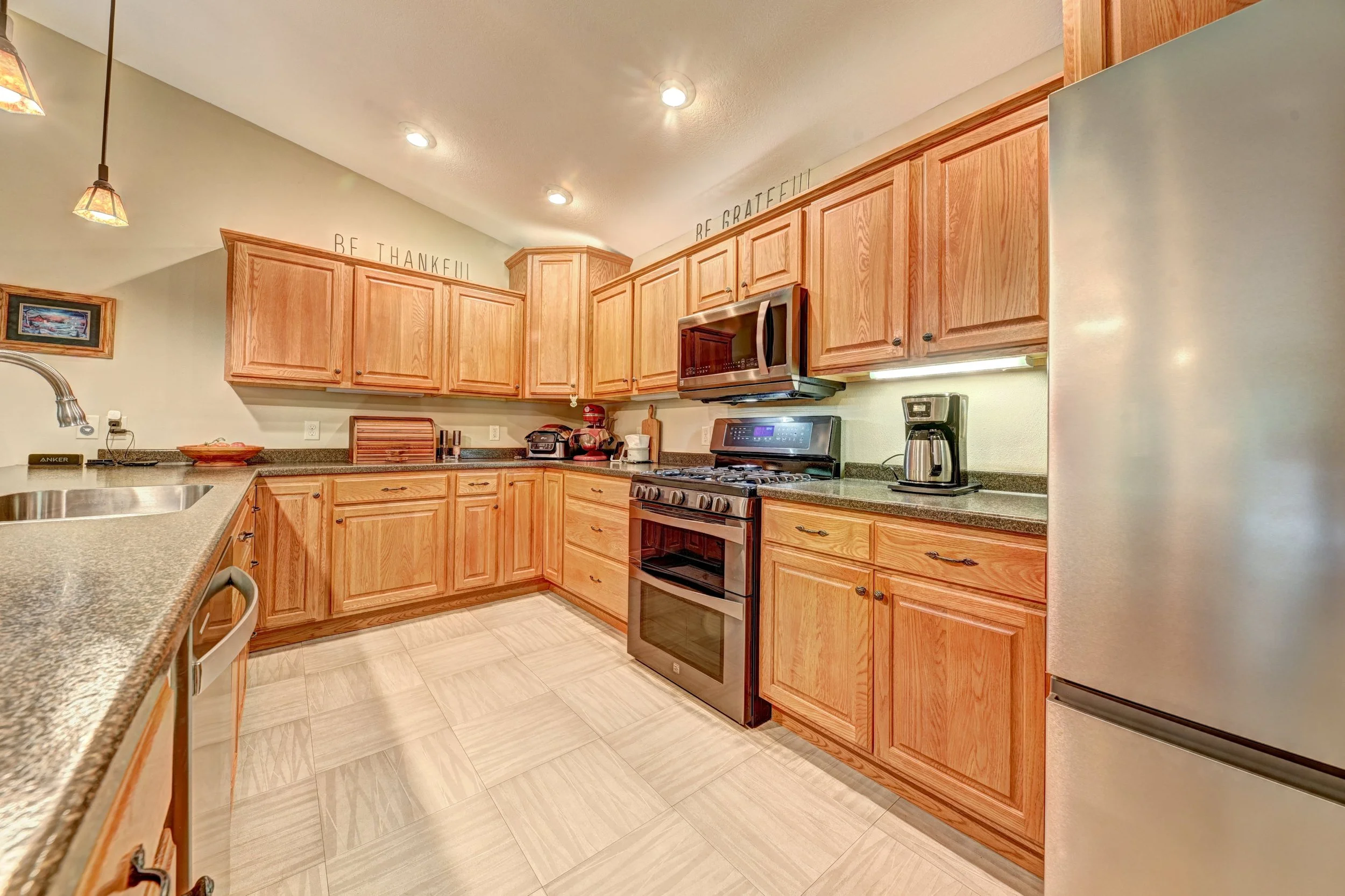 Kitchen with wooden cabinets, granite countertops, stainless steel appliances including a stove, microwave, coffee maker, and dishwasher. Recessed lighting in the ceiling, a sink, and decorative wall signs that say 'Be Thankful' and 'Be Grateful.'