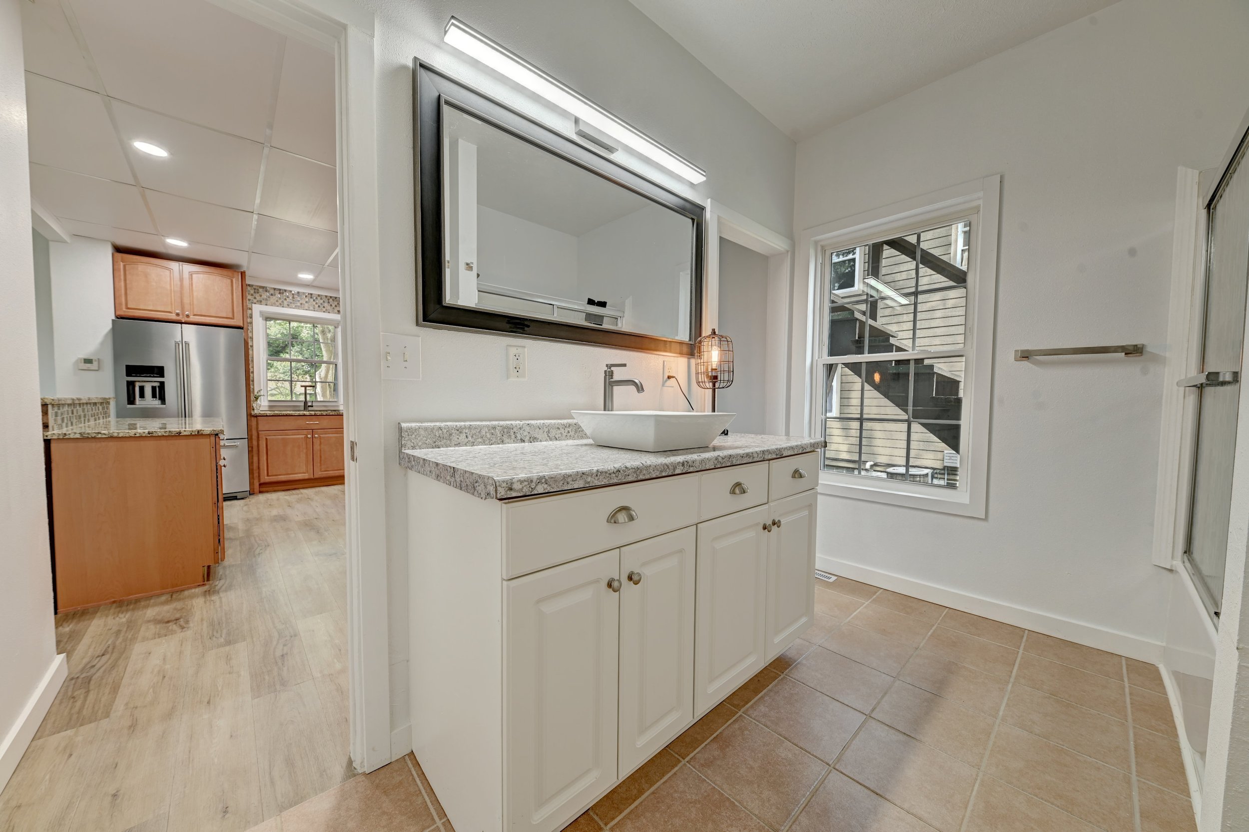 A kitchen with a white cabinet and a vessel sink, light stone countertop, two large windows, and a sliding glass door leading outside. Wood flooring and a view into another kitchen area with wood cabinets and stainless steel refrigerator.