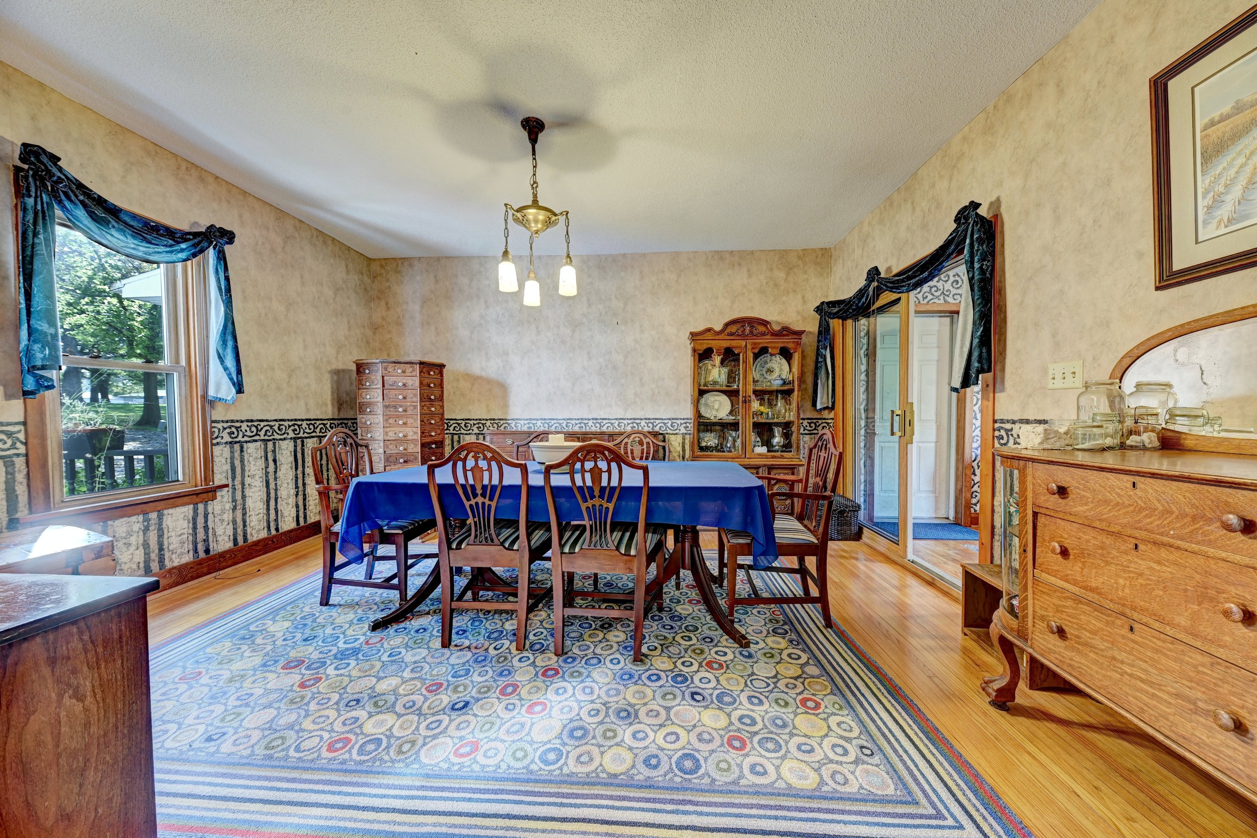 A dining room with a round table covered with a blue tablecloth, wooden chairs, a wooden cabinet with decorative plates, windows with blue curtains, a chandelier, framed pictures, and a patterned carpet.