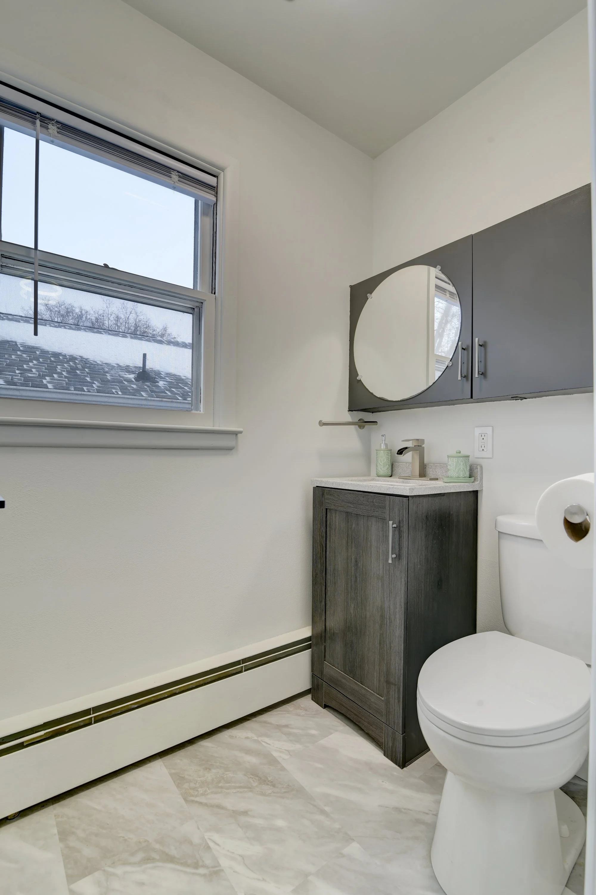 Small bathroom with a window, gray cabinetry, a round mirror, a single sink, and a toilet.