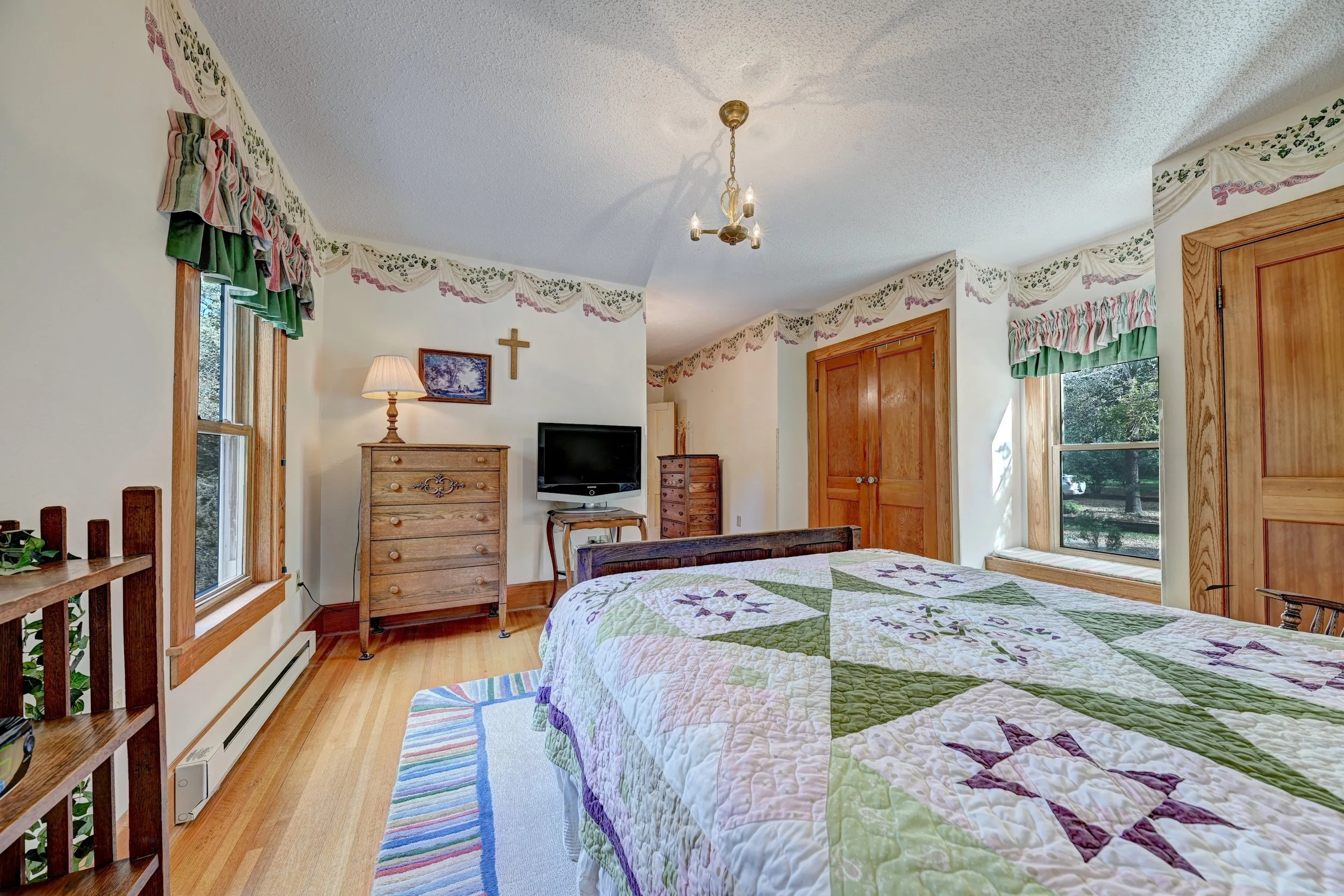 A cozy bedroom with two twin beds with patchwork quilts. The room has two windows with scalloped valances, a small wooden dresser, a TV, a table lamp, and a wall decoration of a cross. There is a colorful striped rug on the wooden floor.