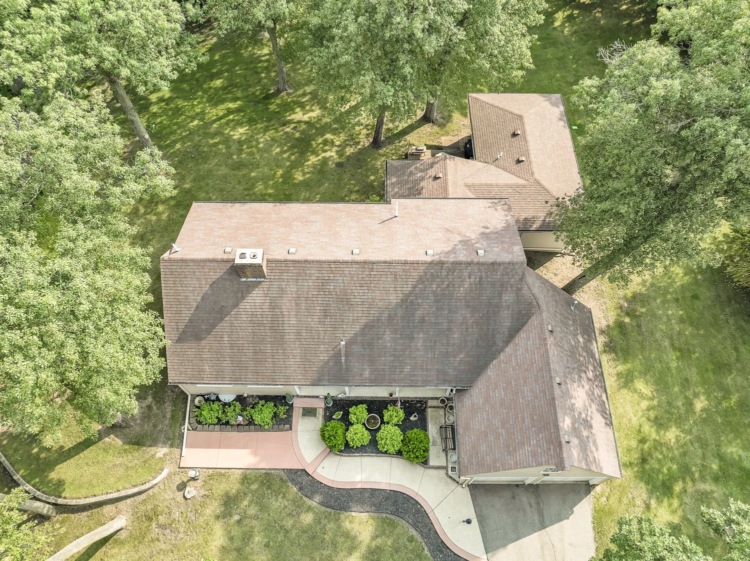 Aerial view of an in-town acreage home with a brown roof, surrounded by green trees and grass, with a curved sidewalk and small landscaped garden area in the backyard.