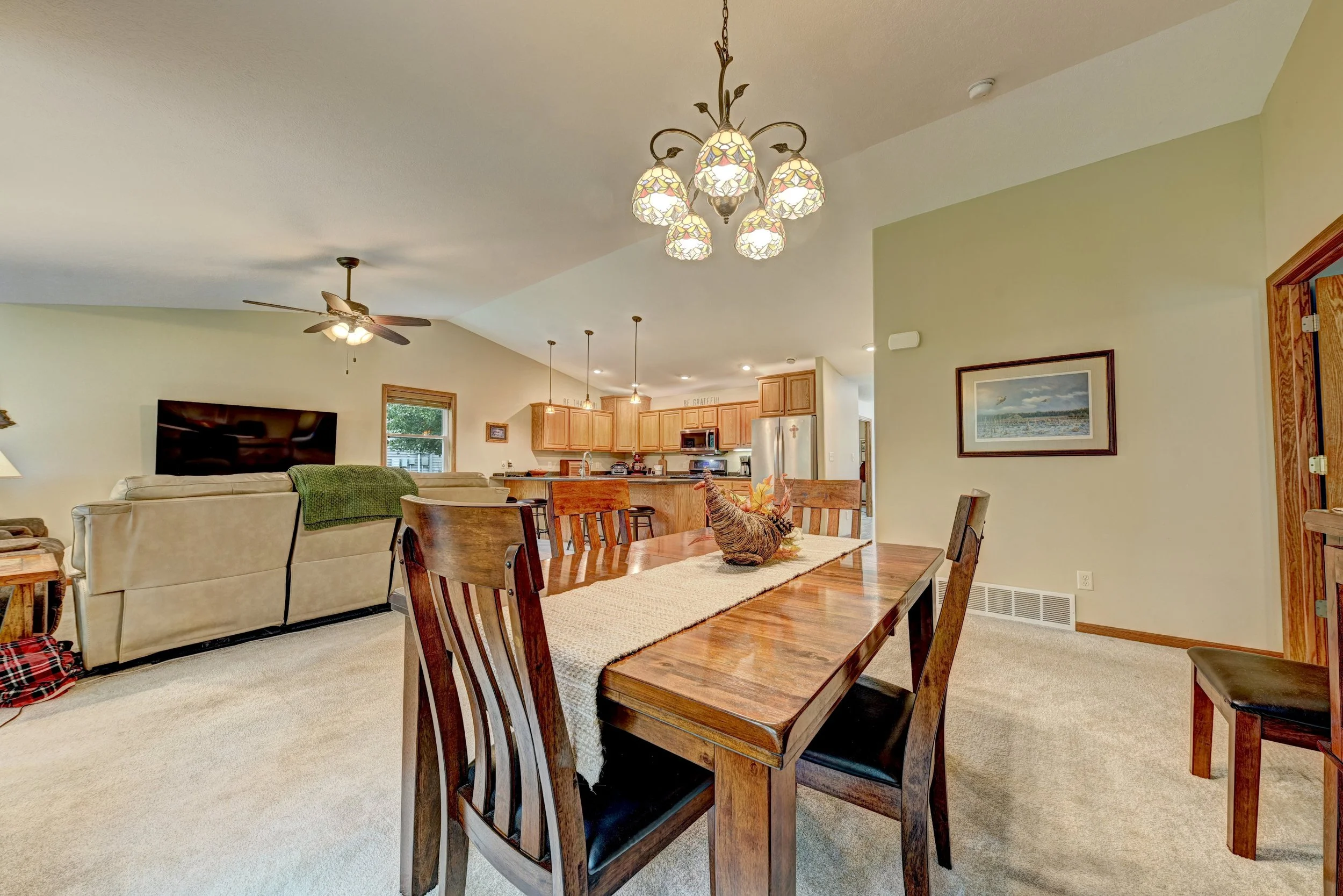 Interior view of a living and dining area with a wooden dining table and chairs, a sofa with a green blanket, a ceiling fan, chandelier, kitchen in the background, framed artwork on the wall, and a window with blinds.