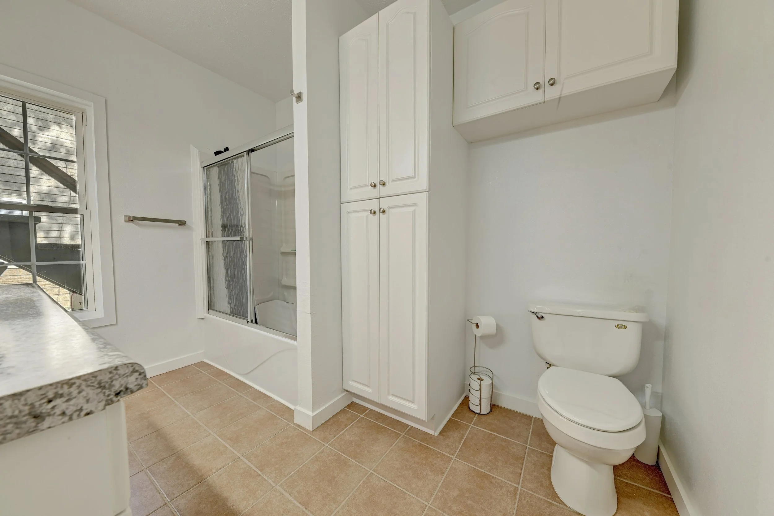 A bathroom with a toilet, white cabinets, a shower with a sliding glass door, a window with blinds, and tiled floor.