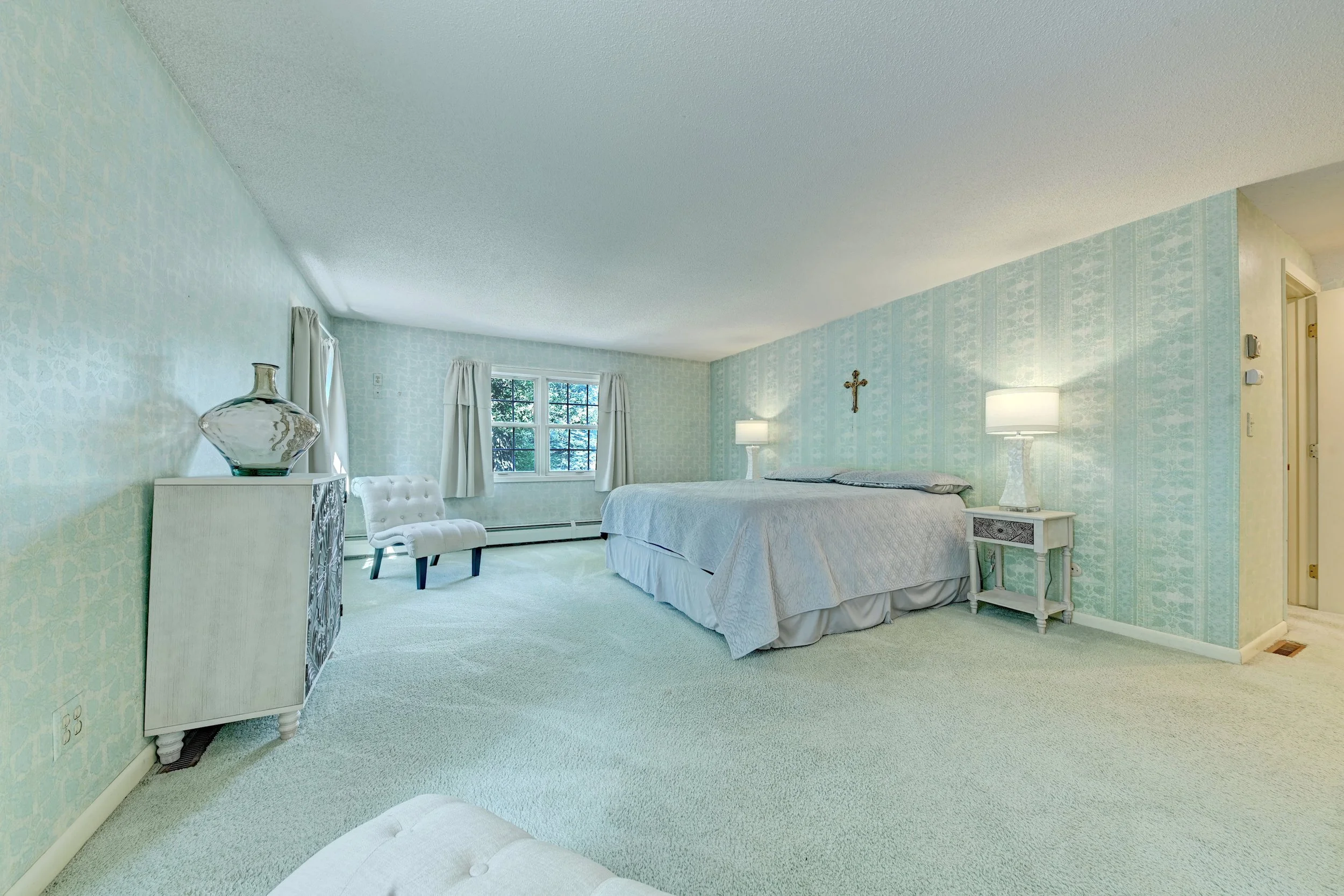 A bedroom with a bed, nightstand, lamps, decorative cross on the wall, window with curtains, a chair, and a glass vase on a dresser, light green wallpaper and carpet.