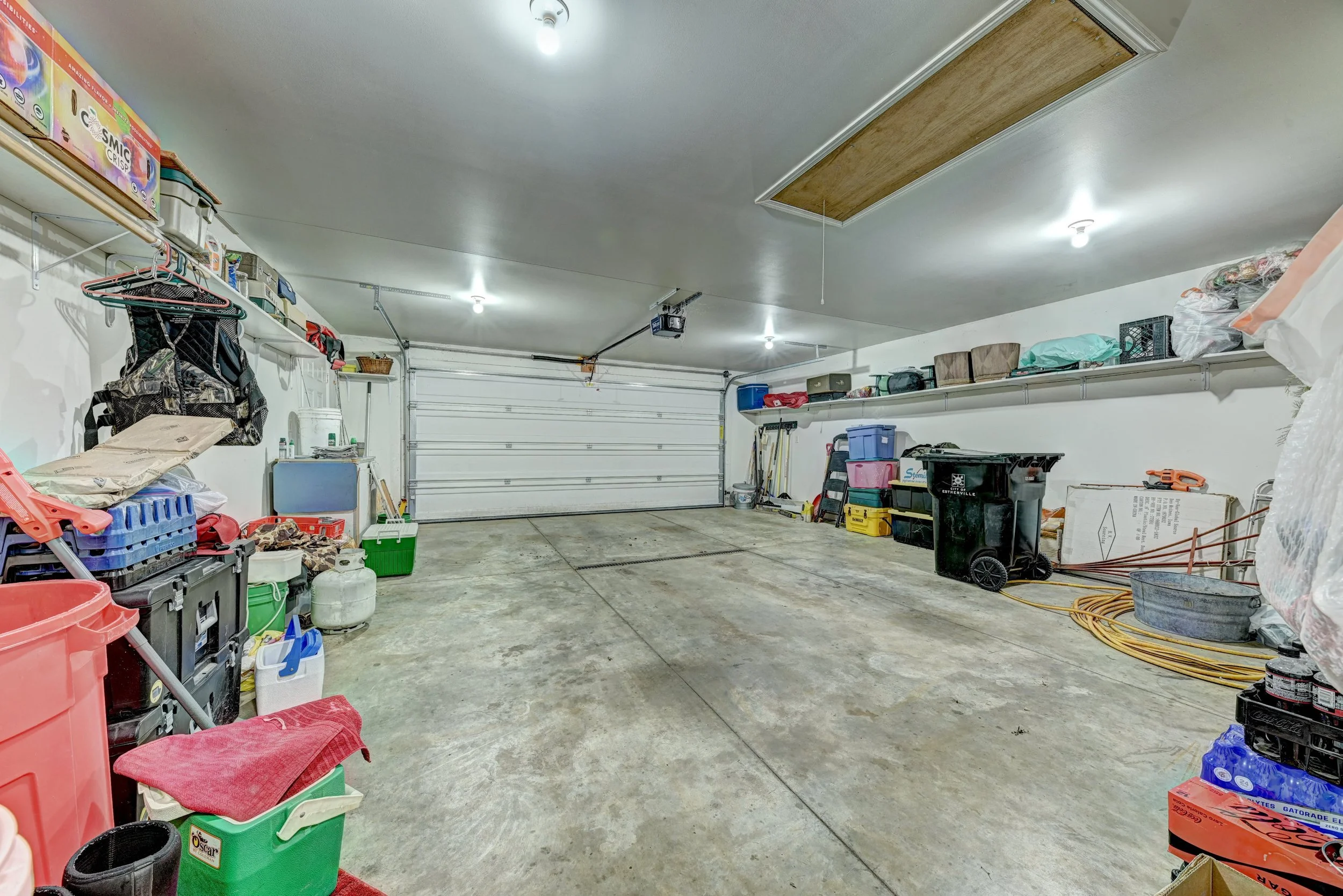 A garage with a concrete floor, white walls, and a white roll-up door. Shelves with various boxed items and storage bins line the walls. There are tools, gardening supplies, hoses, and other miscellaneous items stored neatly on the shelves and on the