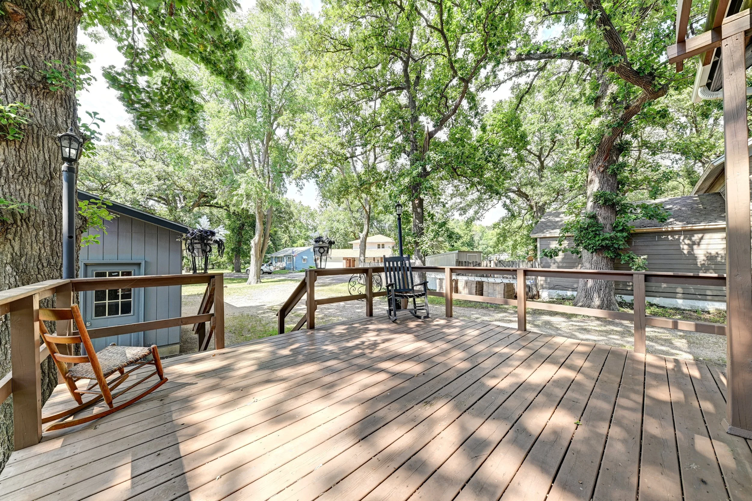 A backyard deck with outdoor chairs, overlooking a yard with trees and houses in the background, shaded by large green leafy trees.