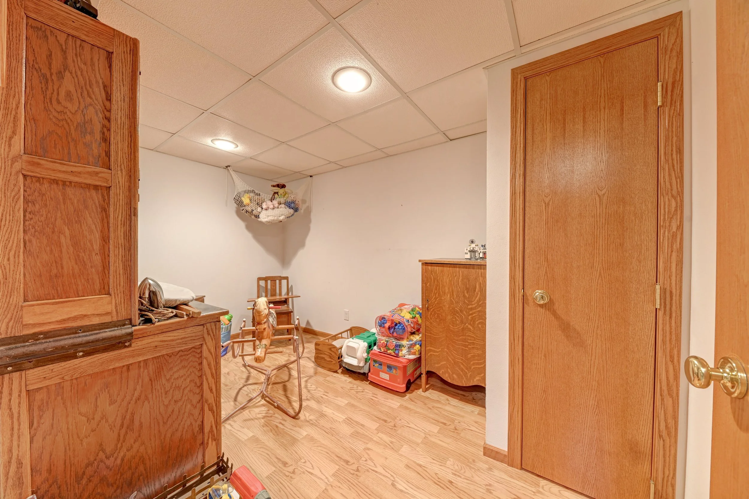 View of a basement room with wood paneling and white ceiling tiles, containing children's toys, a small chair, and a hanging mesh storage bag.