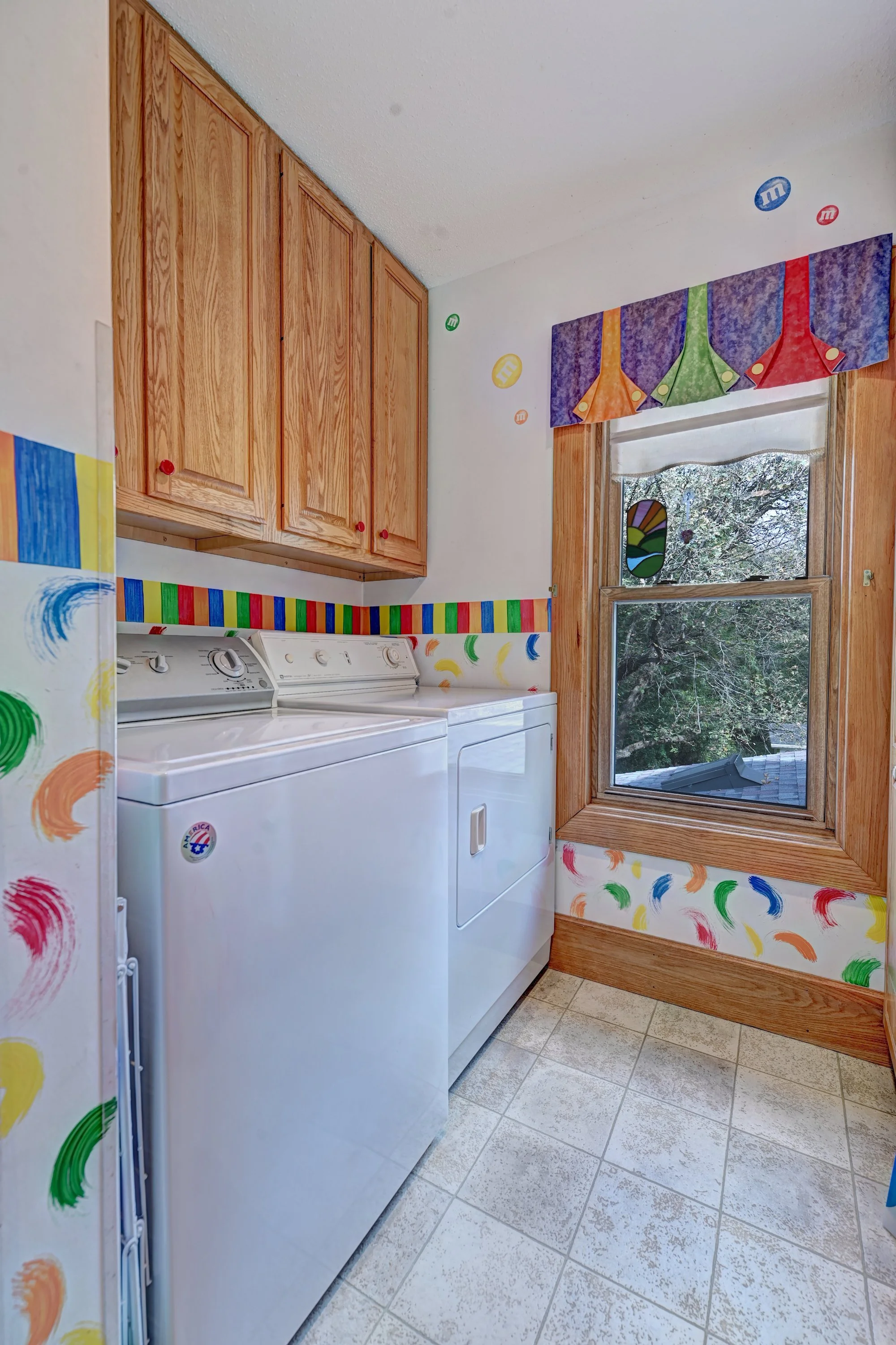 A laundry room with wooden cabinets, a white washing machine and dryer, and a window showing trees outside, decorated with colorful M&M's and swirls on the walls and window valance.