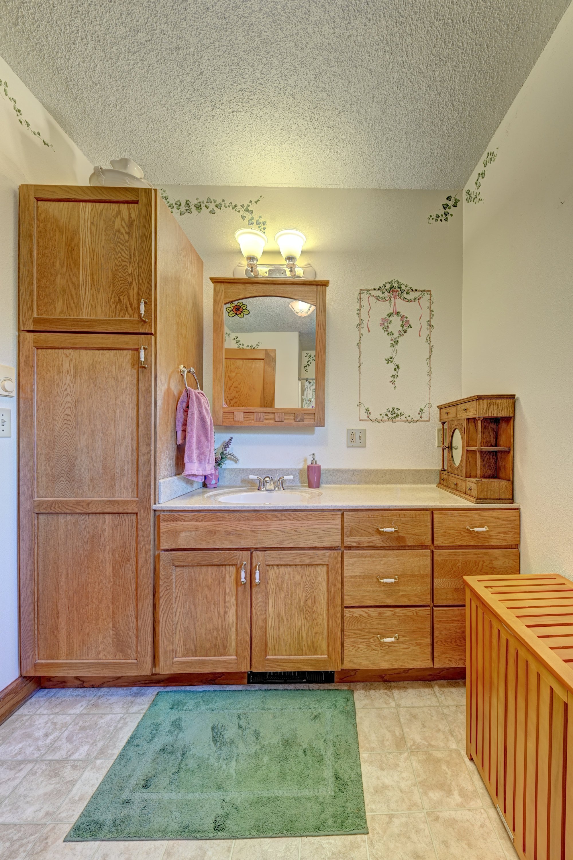 A bathroom vanity area with wooden cabinets, a mirror, a pink towel hanging on a hook, green and beige rugs on tile floor, and decorative wall art featuring green vines.