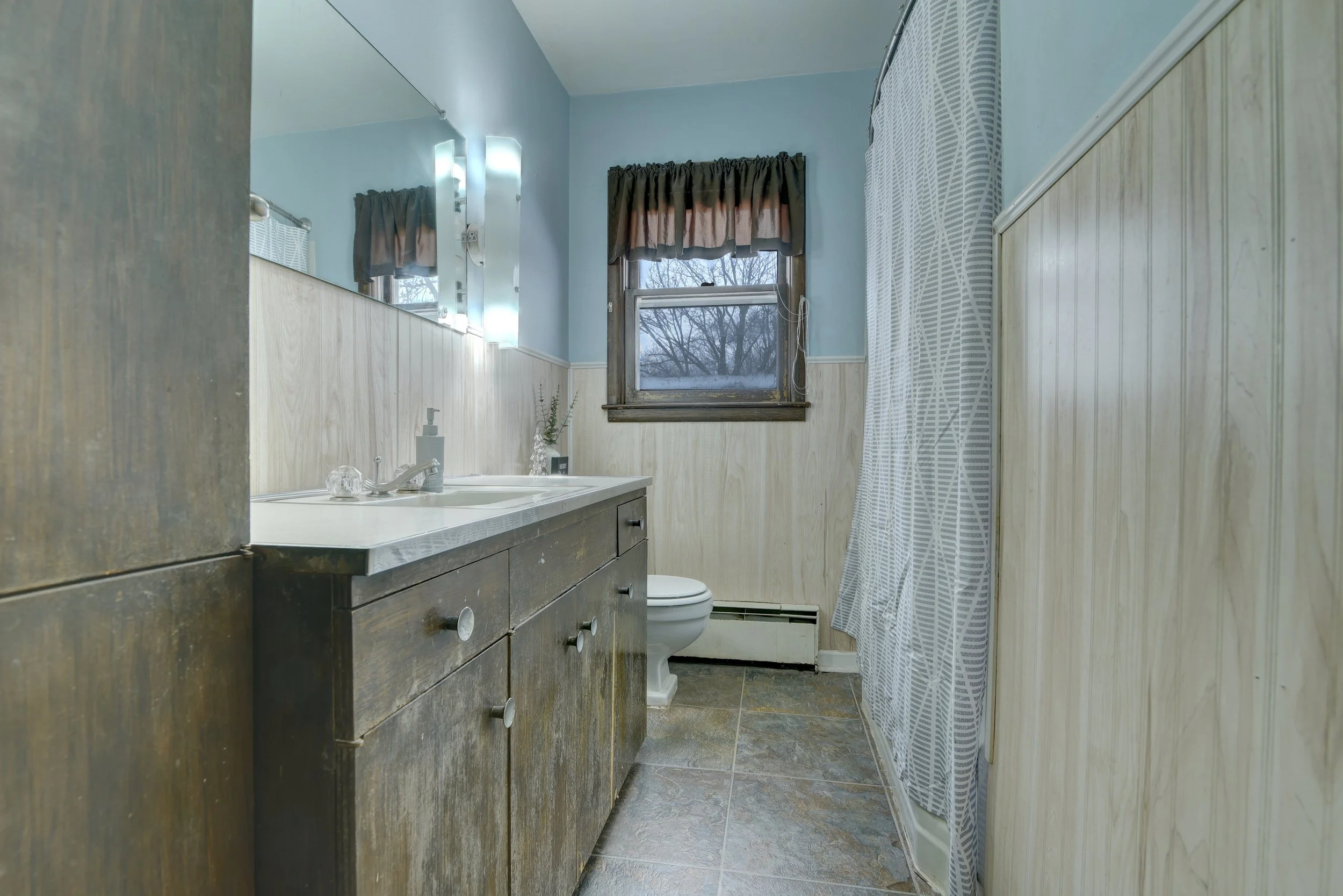 Bathroom with wooden vanity, mirror with light fixtures, window with brown and pink valance, shower curtain with leaf pattern, and tiled floor.