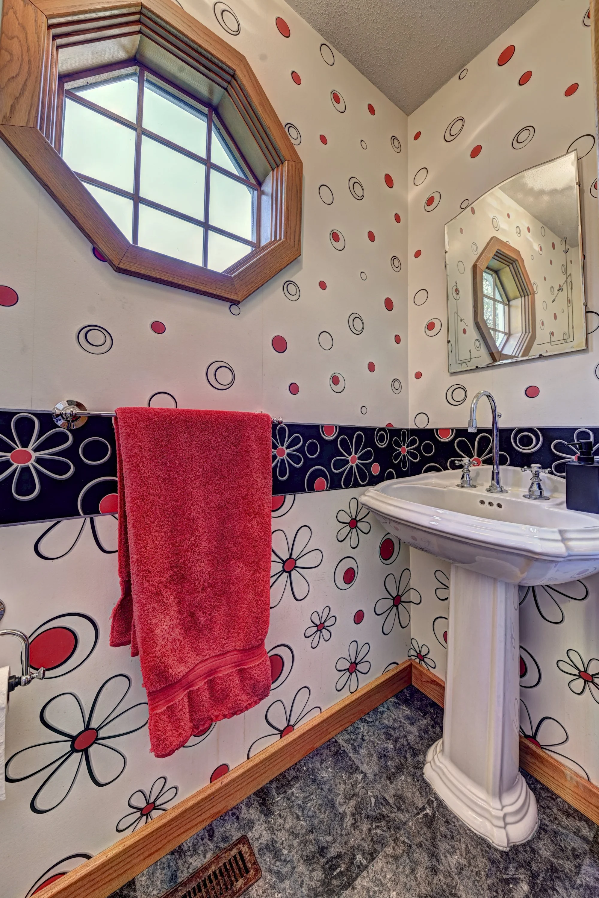 A bathroom with white walls decorated with black and red circle and flower patterns. A window with a wooden frame is in the corner, and a mirror reflects its design. A white pedestal sink with silver fixtures is below the mirror, with a black soap di