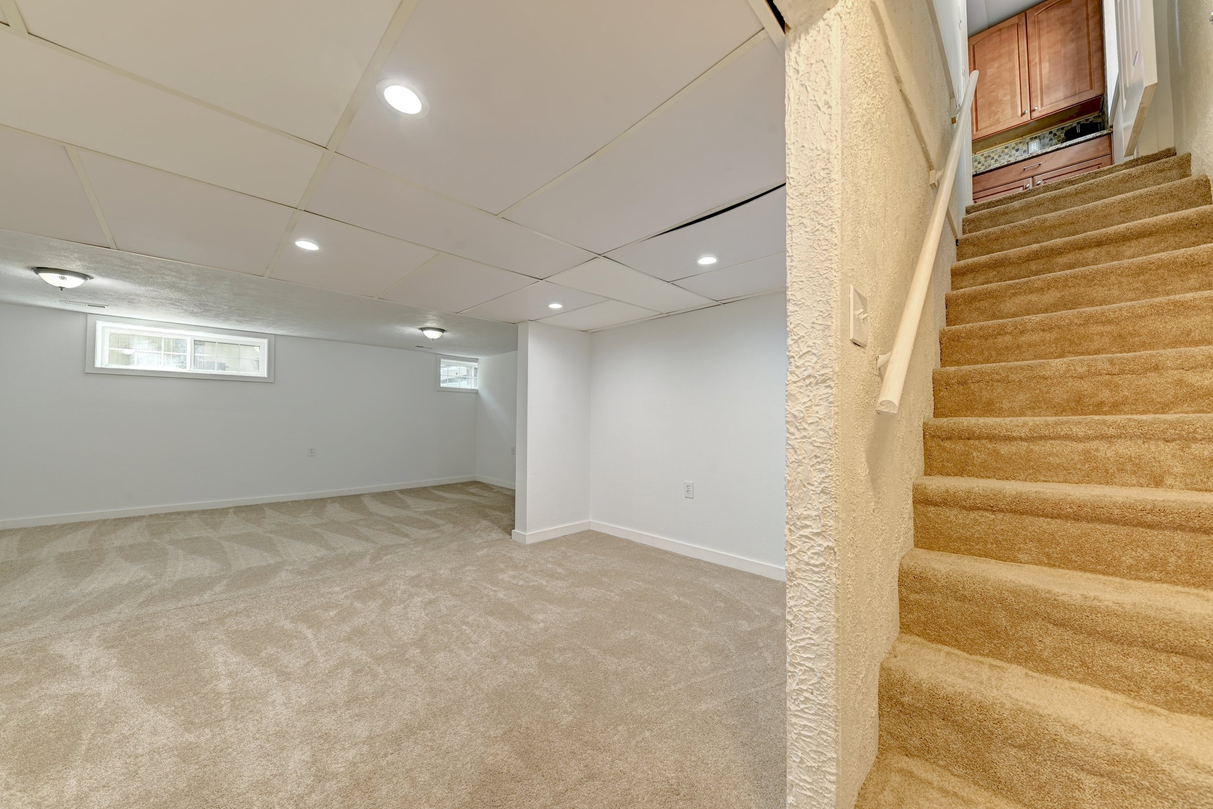 Empty basement room with white walls, beige carpet flooring, and a low rectangular window. There is a staircase with beige carpet leading to an upper level, where wooden cabinets are visible.