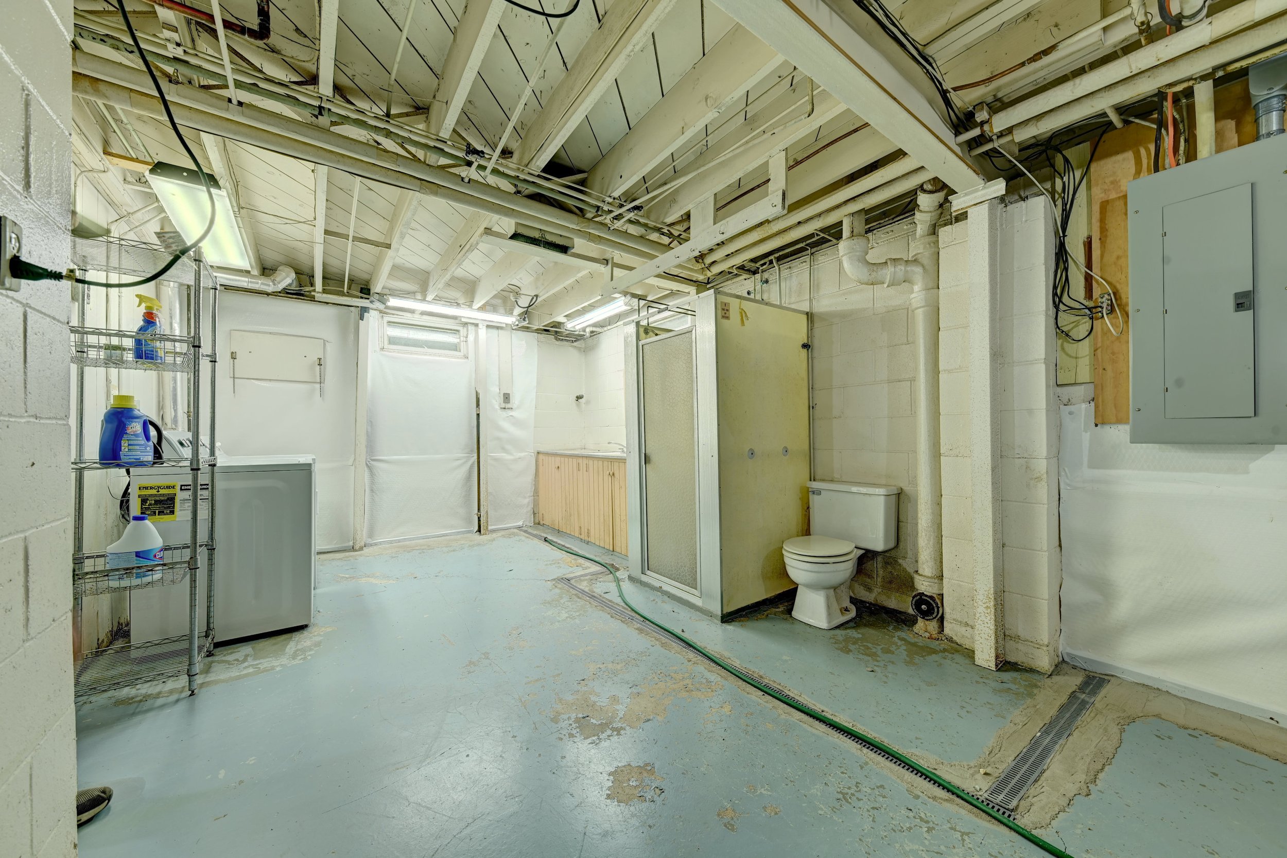 An unfinished basement with exposed ceiling pipes, a toilet, storage shelves with cleaning supplies, and a concrete floor with patches of paint or stains.