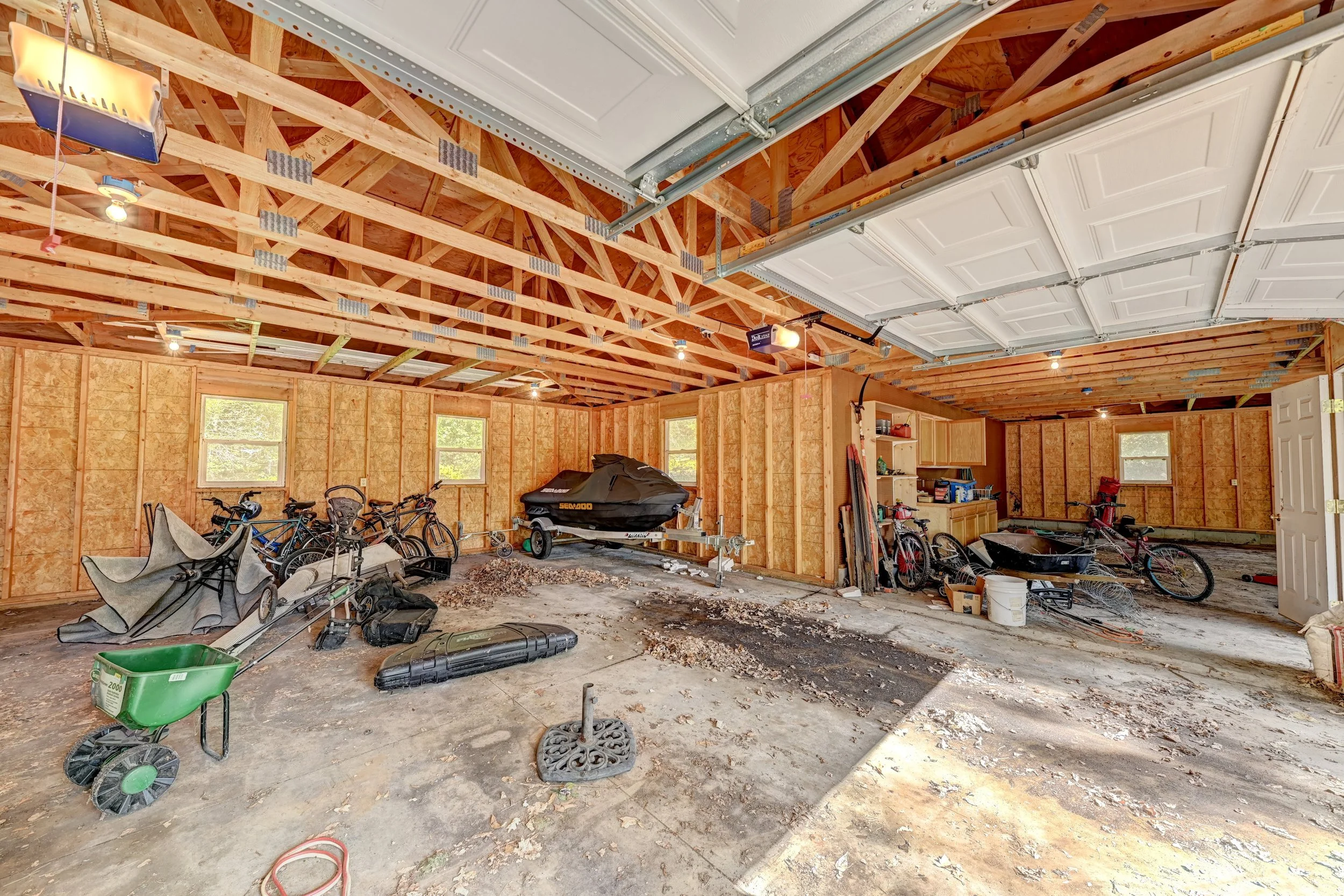 Interior view of an unfinished garage with exposed wooden framing, a partly installed garage door, bikes, a boat on a trailer, and various tools and equipment scattered across the floor.