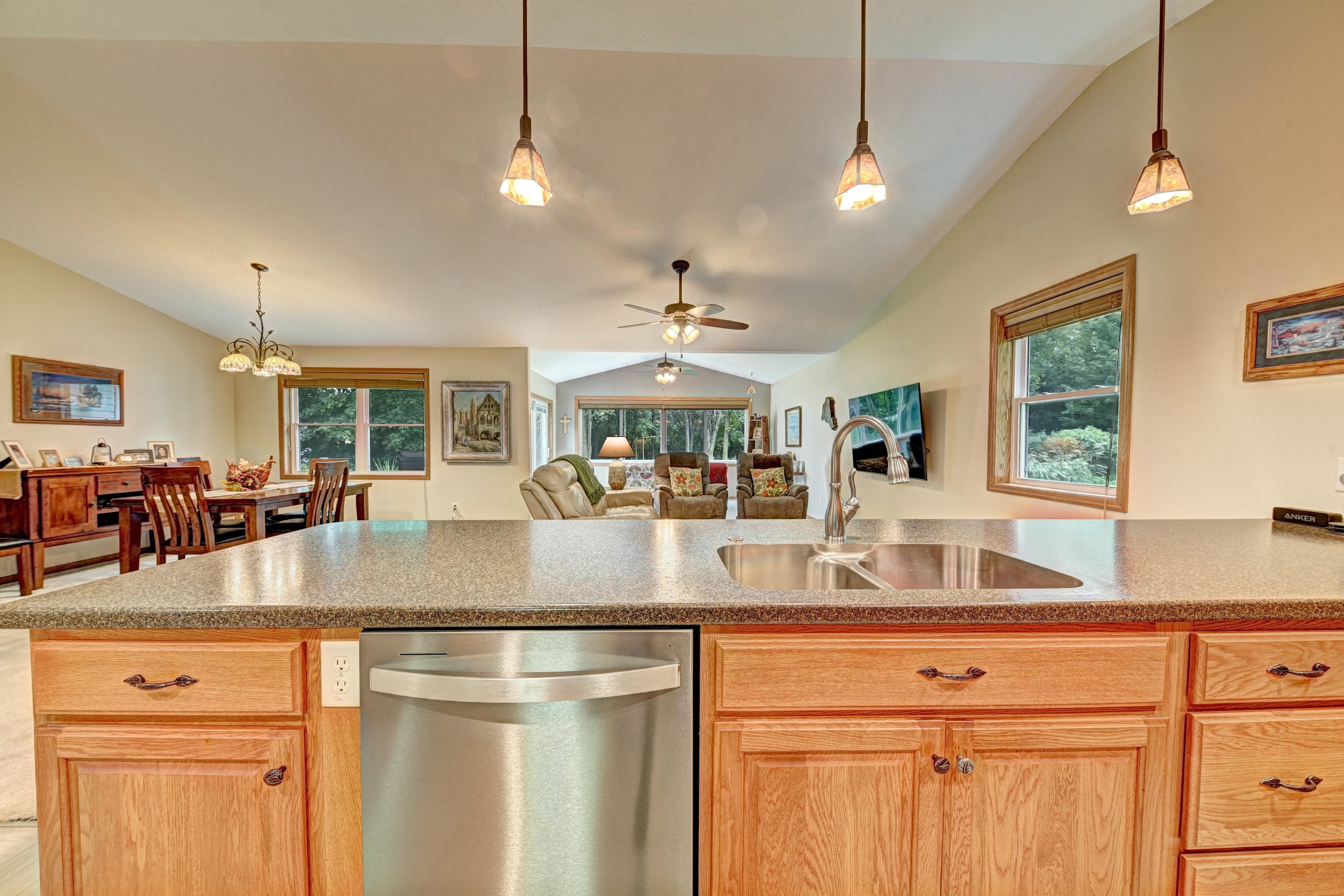 A kitchen view looking into a living room, with a granite countertop and wooden cabinets, stainless steel dishwasher, and a kitchen sink. The living room features beige sofas, armchairs with floral pillows, large windows, and ceiling fans with lights
