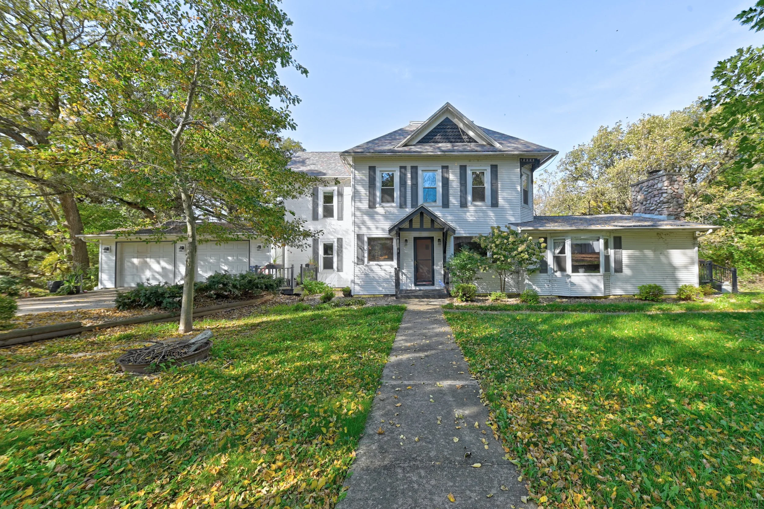 A two-story white house with black shutters, a central porch, and two garage doors on the left side. The house has a steep gabled roof with shingles and a chimney on the right. The front yard has a concrete walkway leading to the front door, surround