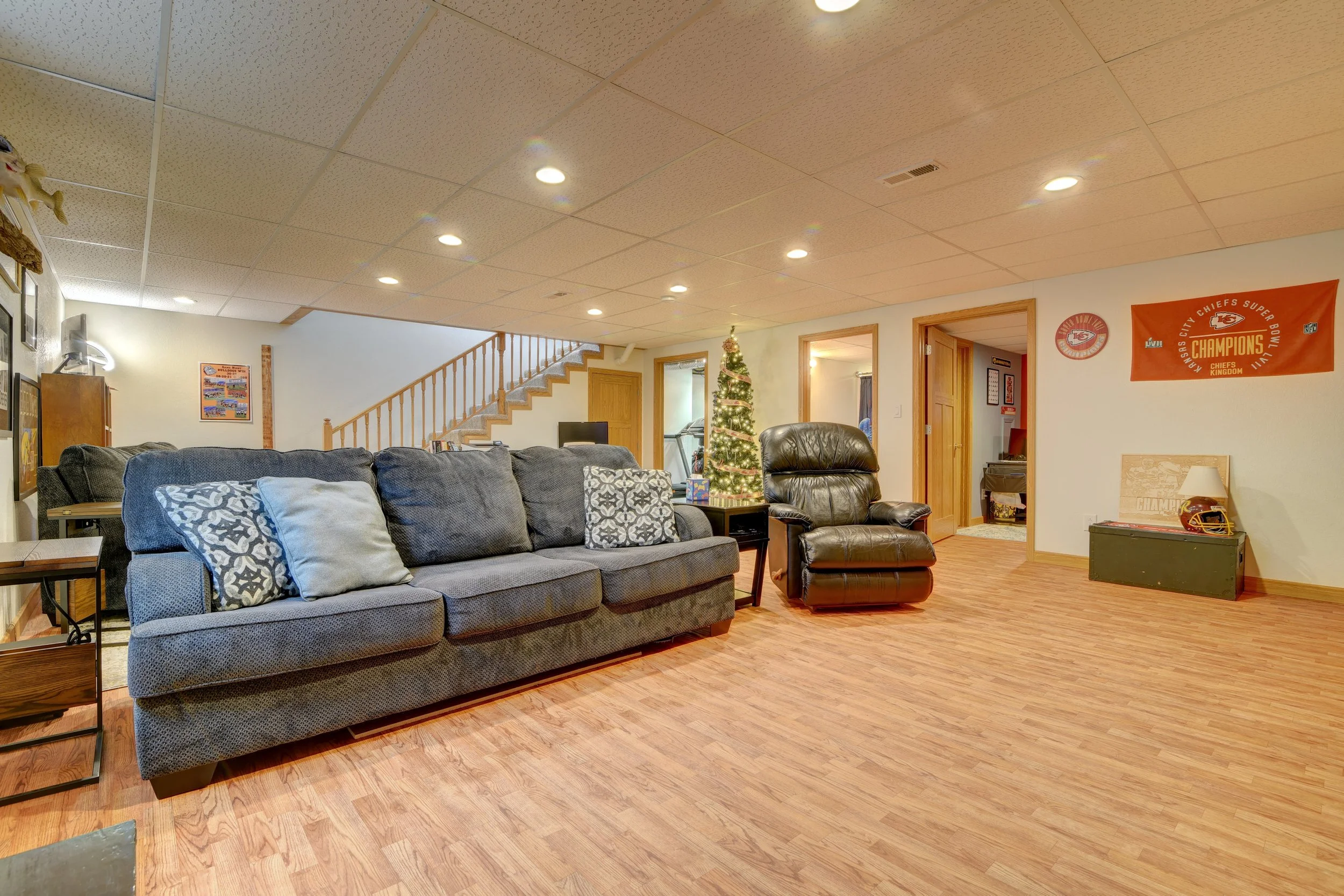 Living room with a gray sofa, a black leather recliner, a decorated Christmas tree, wooden floors, and sports banners on the walls.