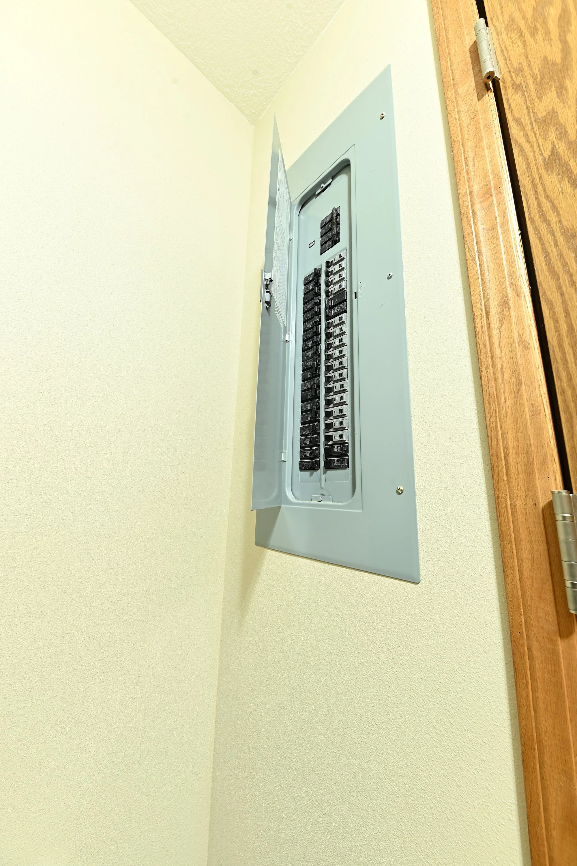 An electrical panel with circuit breakers, mounted on a light yellow wall next to a wooden surface with a hinge.