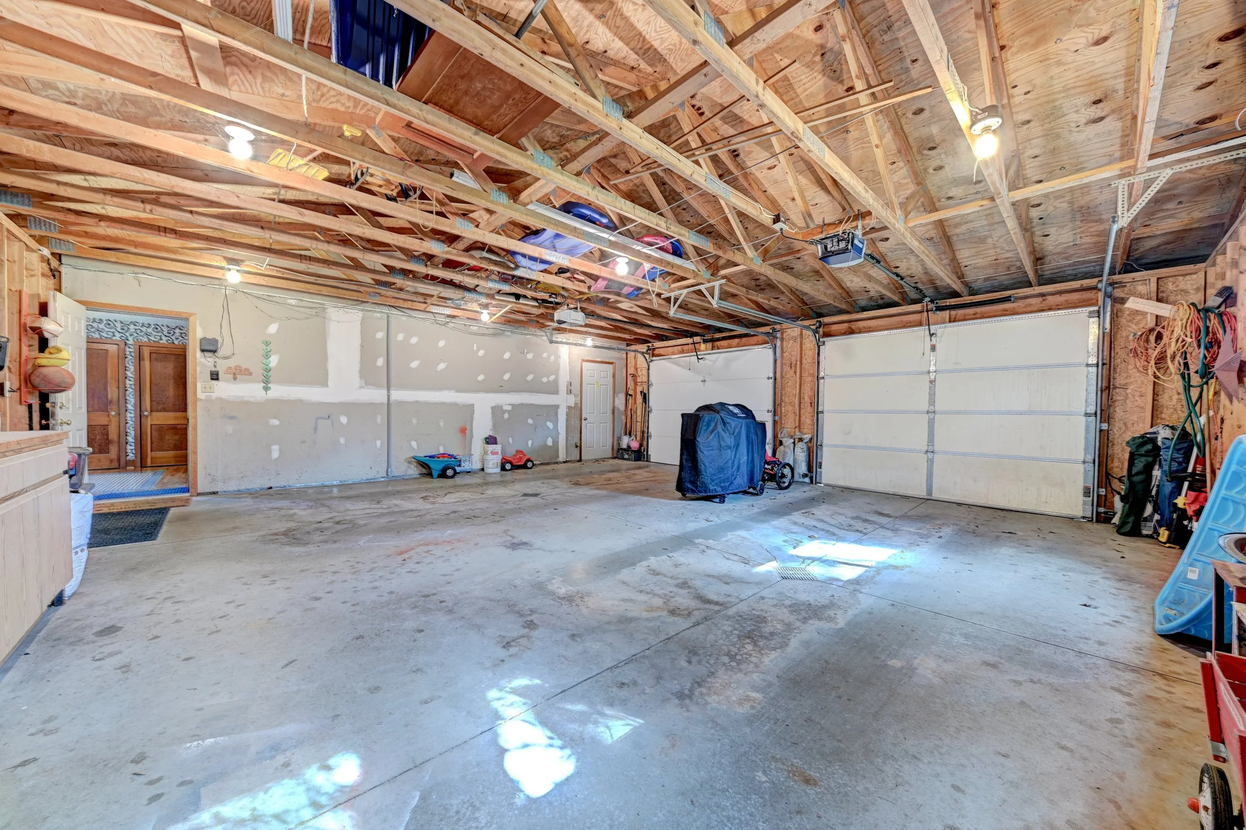 A spacious garage with an unfinished ceiling showing wooden beams and exposed insulation. The garage has a concrete floor and contains various items including a covered object, a toy car, a bike, and some hanging cords and tools. The garage doors are