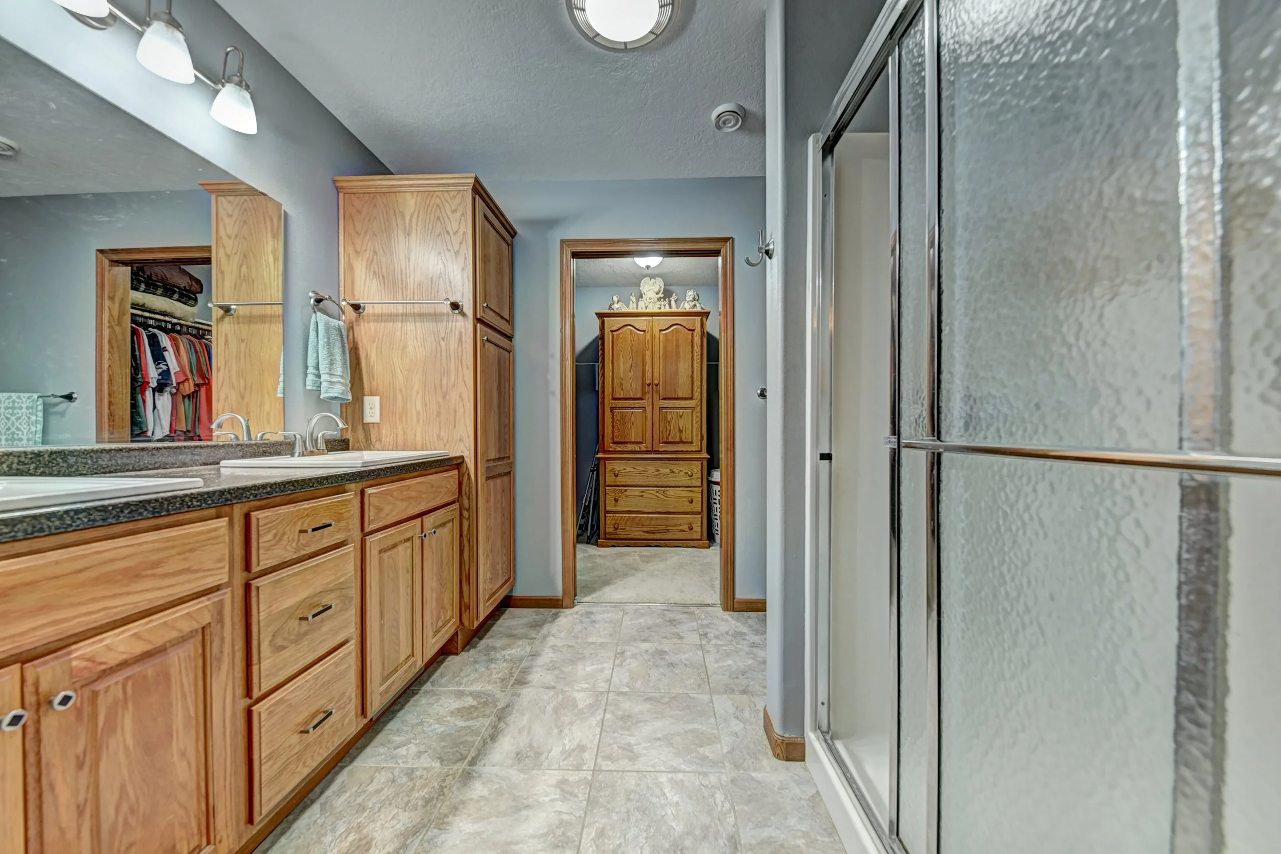 Bathroom with wooden cabinetry, a granite countertop, a double sink, a towel hanging, a bathroom mirror, a washing machine, a shower with glass door, and a closet with clothes inside.